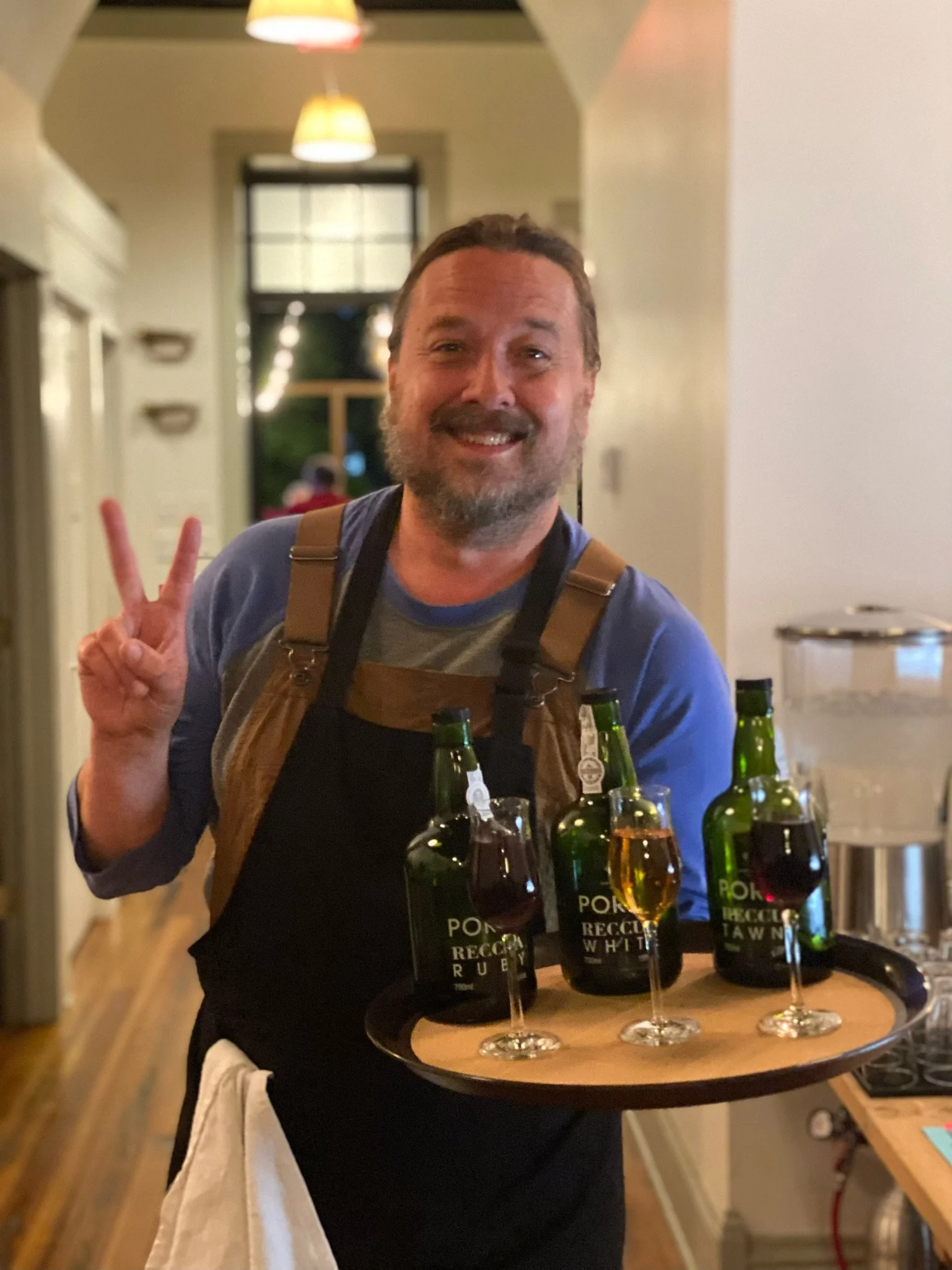 A man with a beard smiling and showing a peace sign, holding a tray with four bottles of beer and four glasses of wine or similar drinks, in a warmly lit indoor setting.