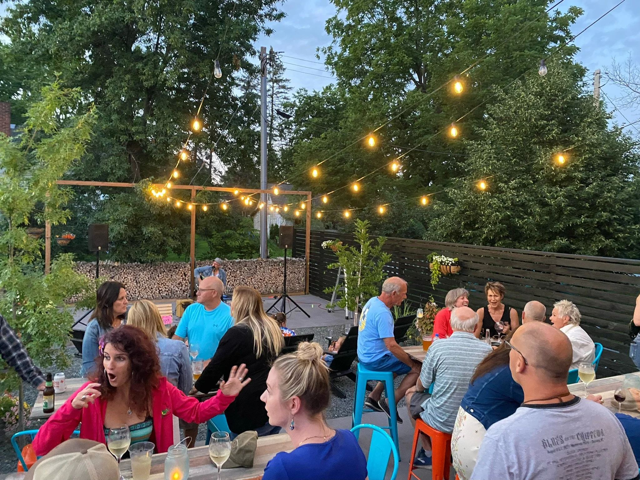 People gather at an outdoor evening party with string lights overhead, a small stage, and a wooden fence in the background. Some are seated at tables with drinks, engaging in conversation.