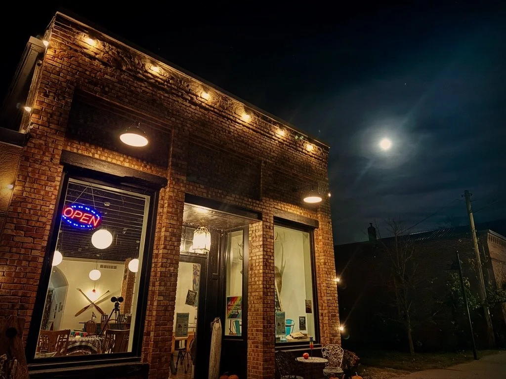 Nighttime view of a brick building with large windows and string lights along the top. An illuminated open sign is visible inside, along with a bicycle and chairs.