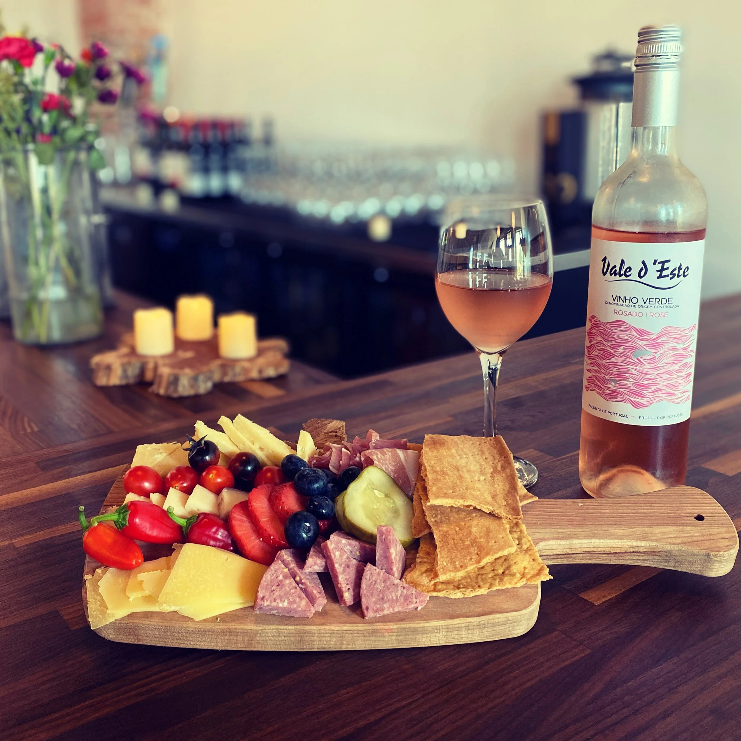 A wooden serving board with cheese, cherry tomatoes, grapes, strawberries, sliced meat, and crackers. A glass of rosé wine and a bottle of rosé wine are on the table. In the background, there are candles and a vase with flowers.