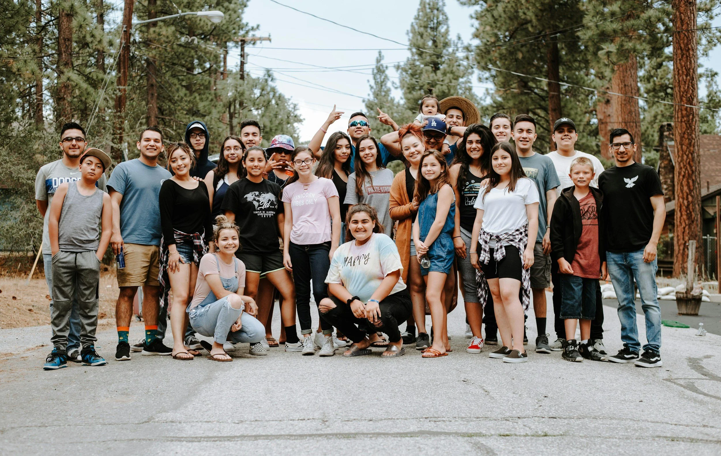 A large group of people, including children and adults, posing together outdoors in a wooded area, with trees, power lines, and a building in the background.