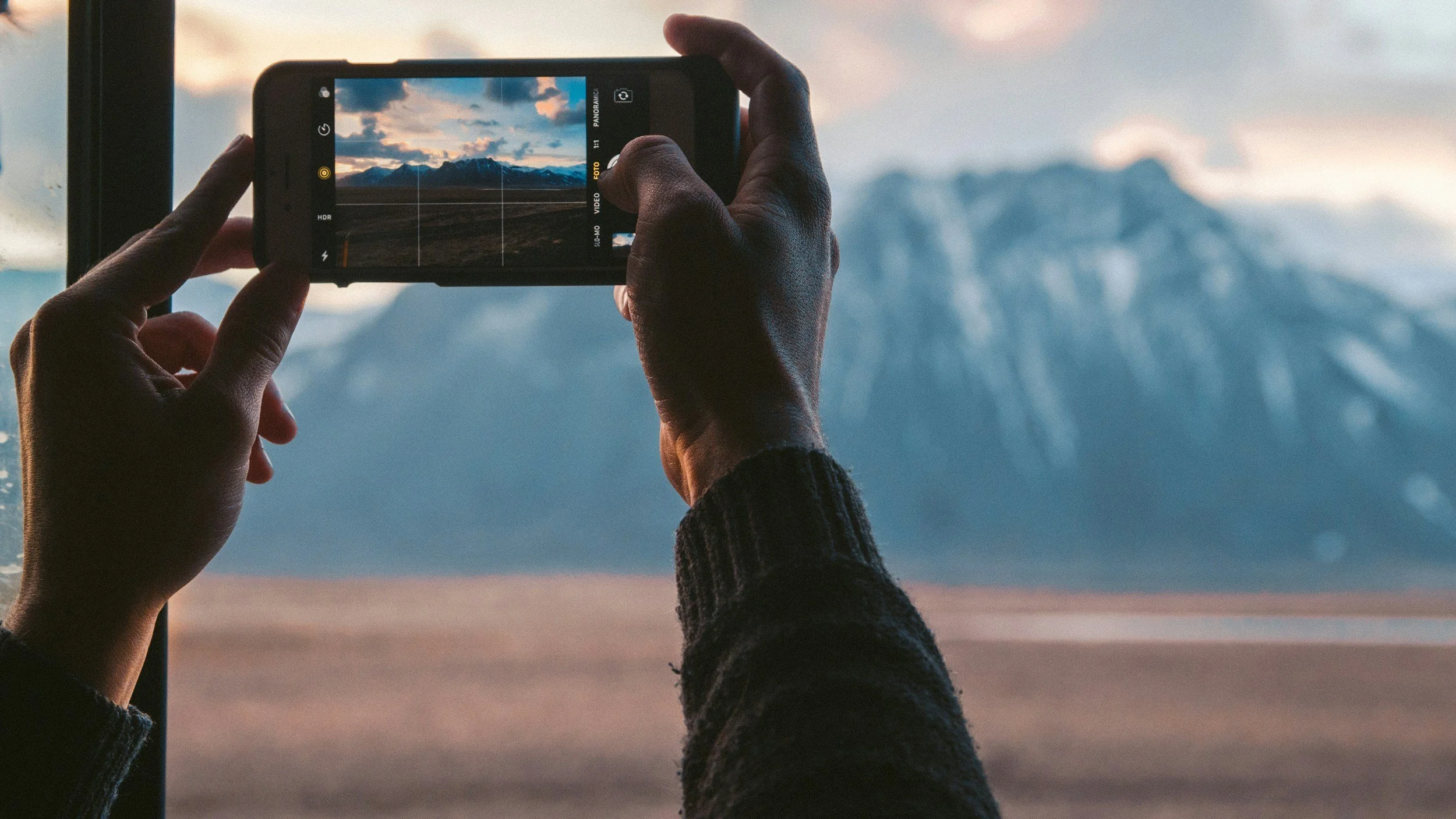 Person taking a photo of a mountain landscape through a window using a smartphone.