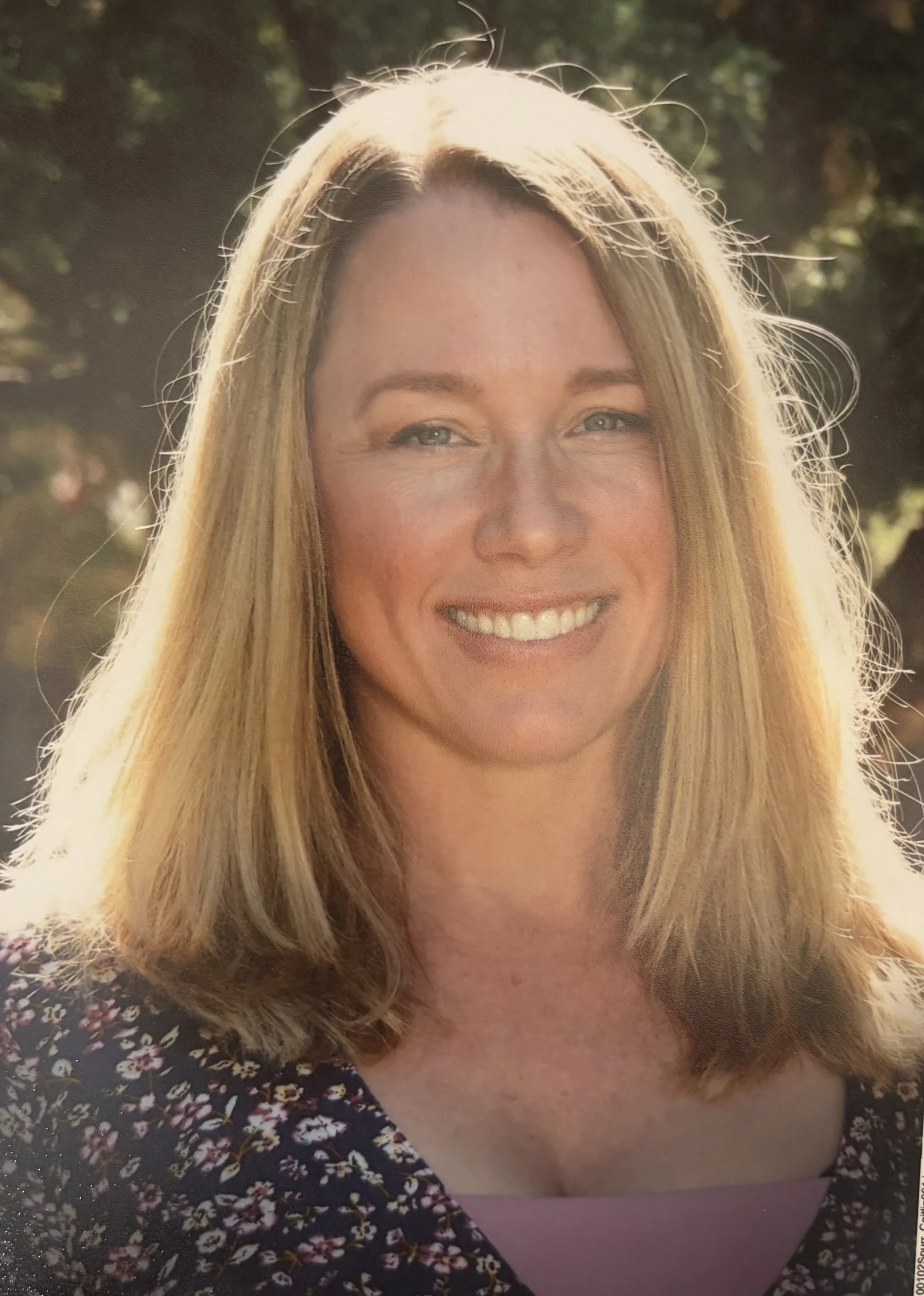 A woman with shoulder-length blonde hair smiling outdoors in sunlight, wearing a floral top with a pink shirt underneath.