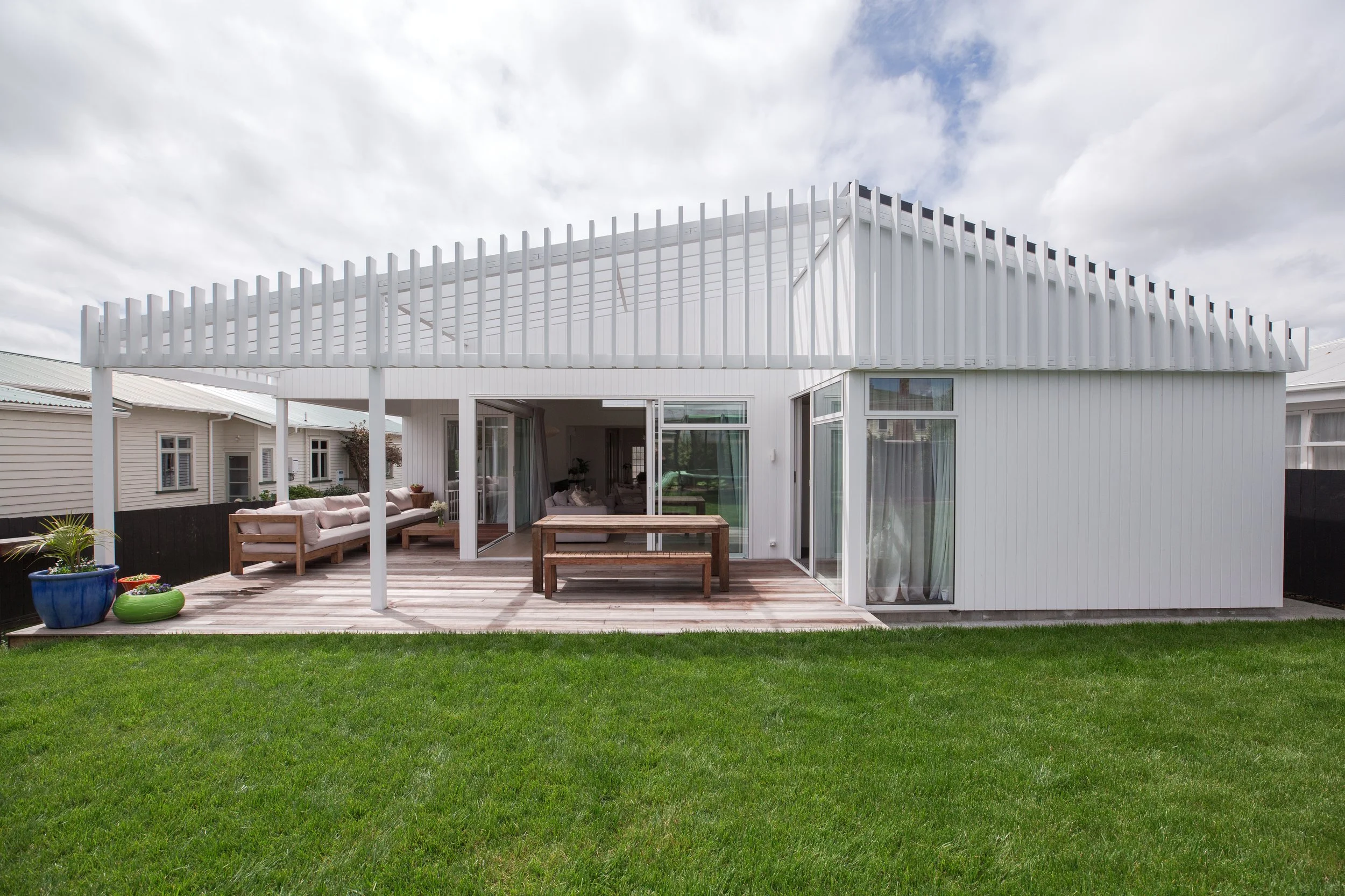 Modern white house with a deck and sliding glass doors, outdoor seating area, and a green lawn, under a partly cloudy sky.