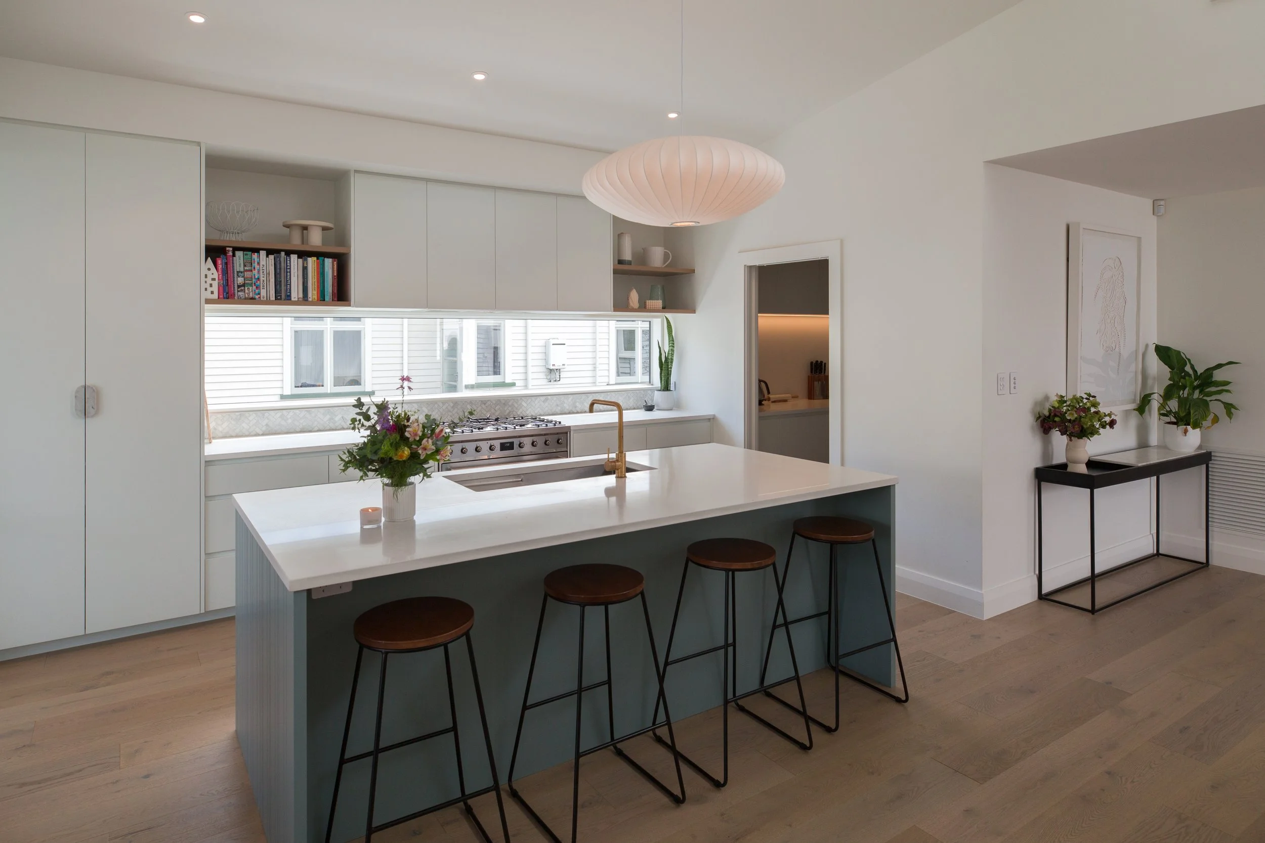 Modern minimalist kitchen with white cabinetry, a kitchen island with a white countertop, four brown bar stools, a gold faucet, a row of windows above the counter, and decorative plants and flowers.