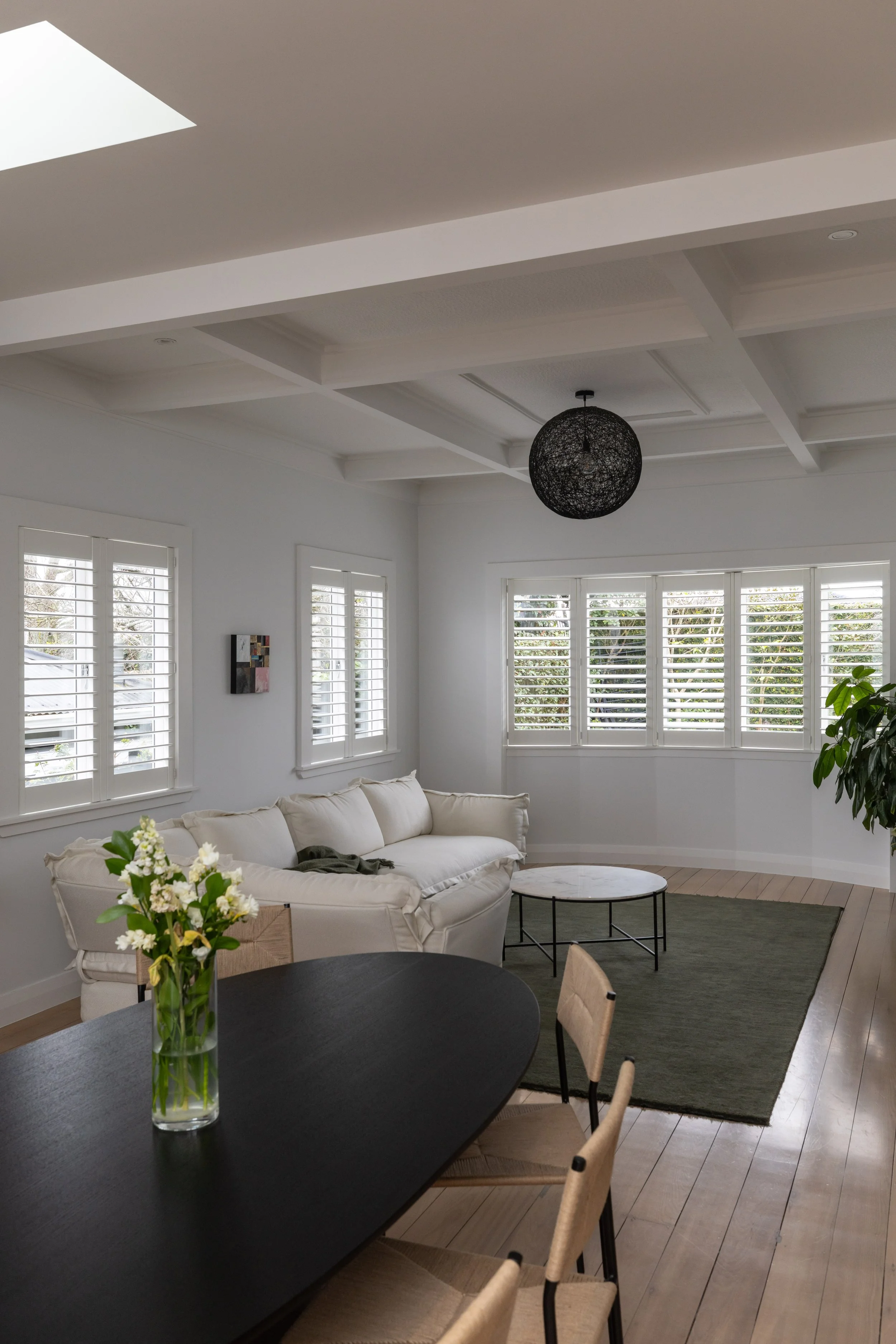 Living room with white walls, white sofa, wooden floor, large bay window with shutters, black pendant light, green area rug, round marble-top coffee table, and a black dining table with beige chairs.