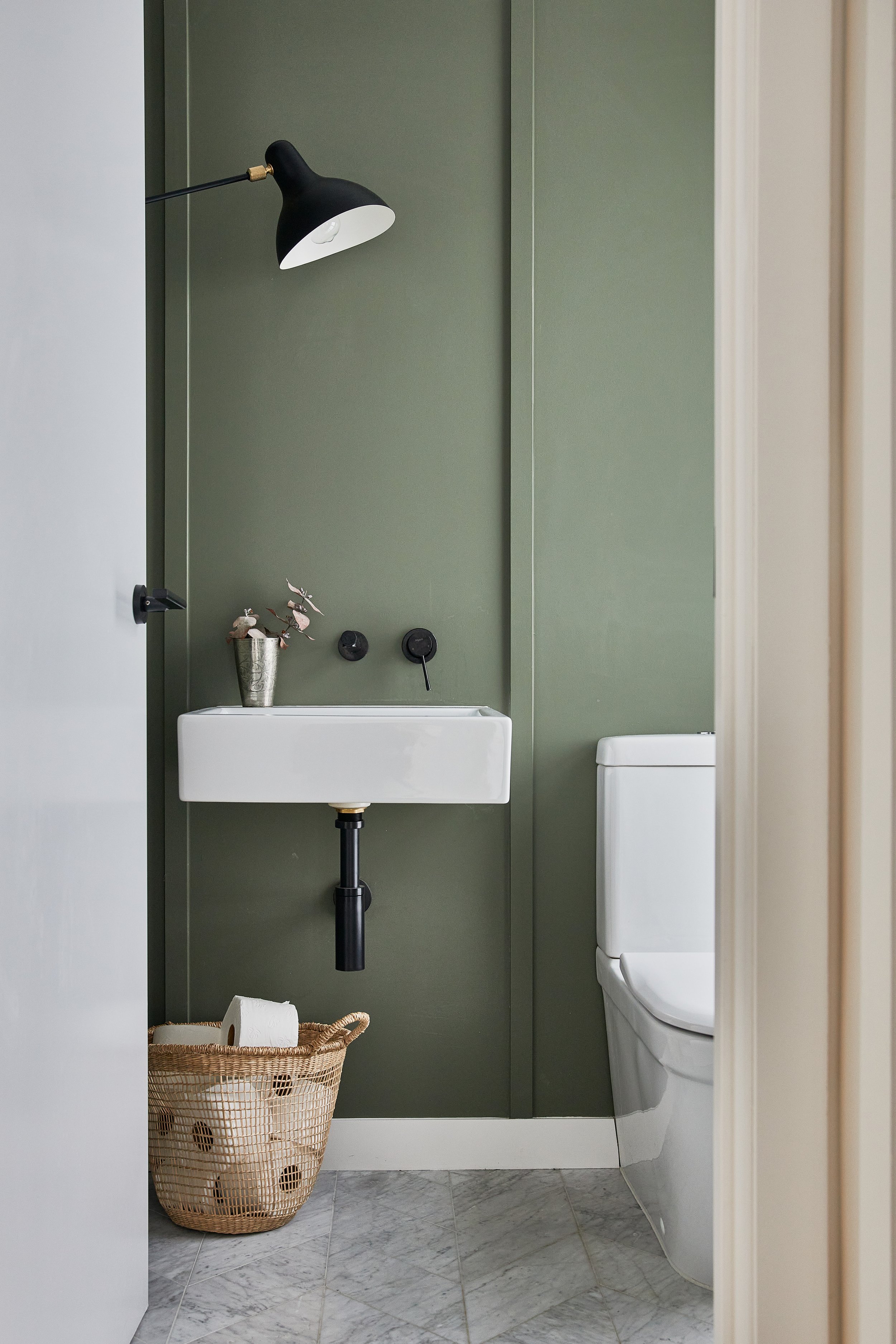 A small bathroom with a green wall, white sink, and white toilet. There is a black wall-mounted light above the sink, a small pink plant in a silver bucket on the sink, and a basket of toilet paper on the floor. The floor is gray marble.