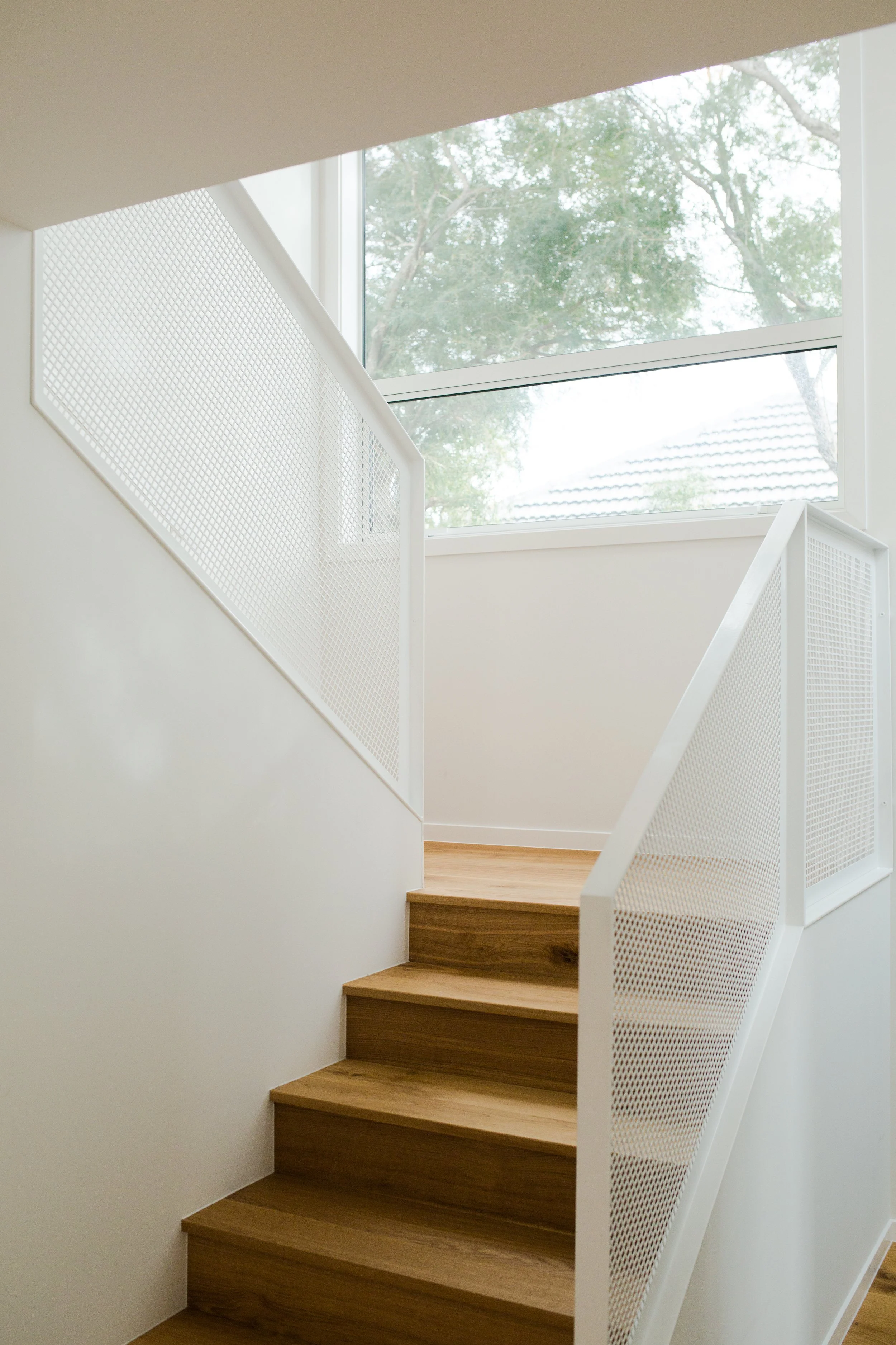 Wooden staircase with white metal railings in a bright room with large windows showing trees outside.