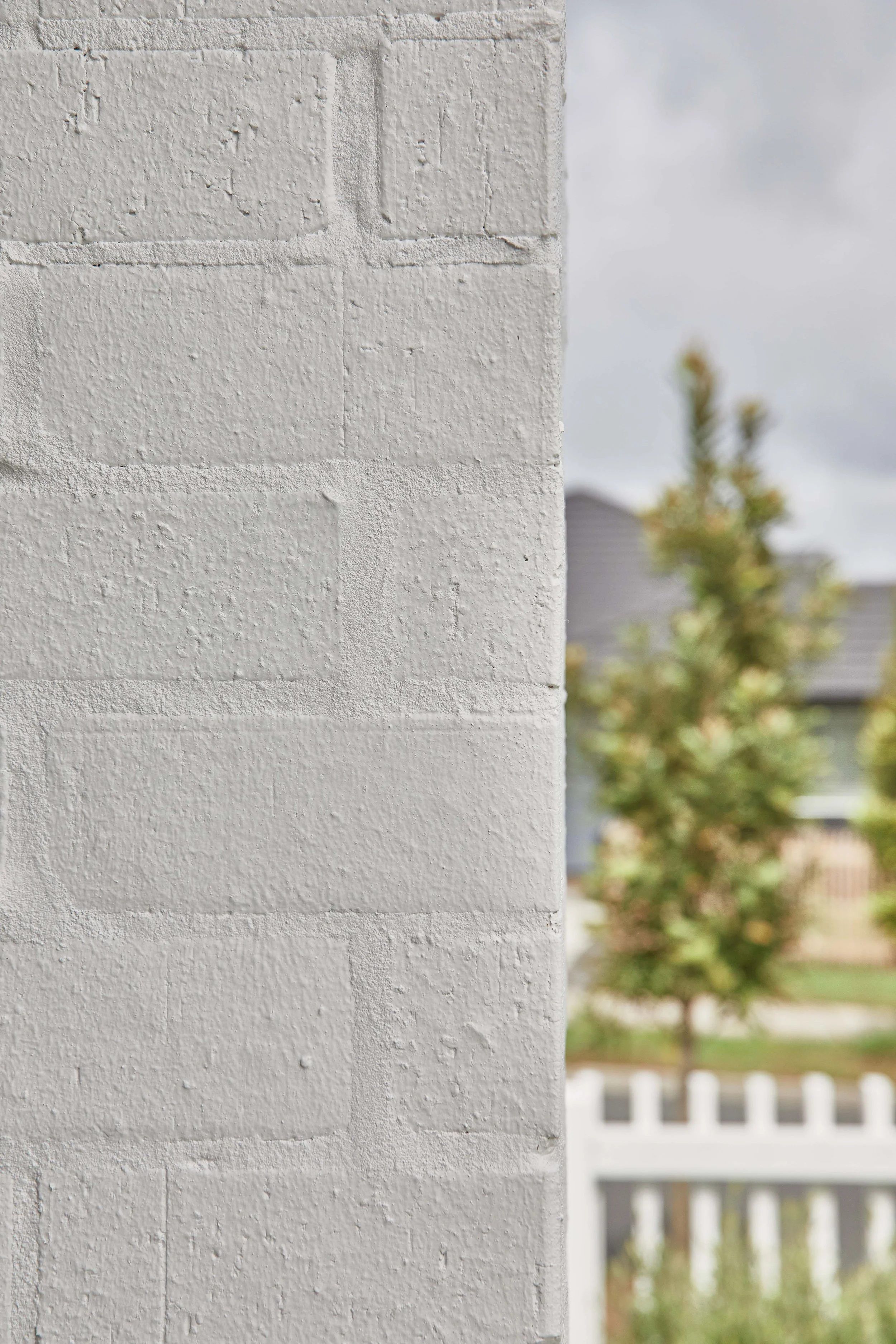 Close-up of a white brick wall with a blurred background of trees, houses, and a white picket fence.