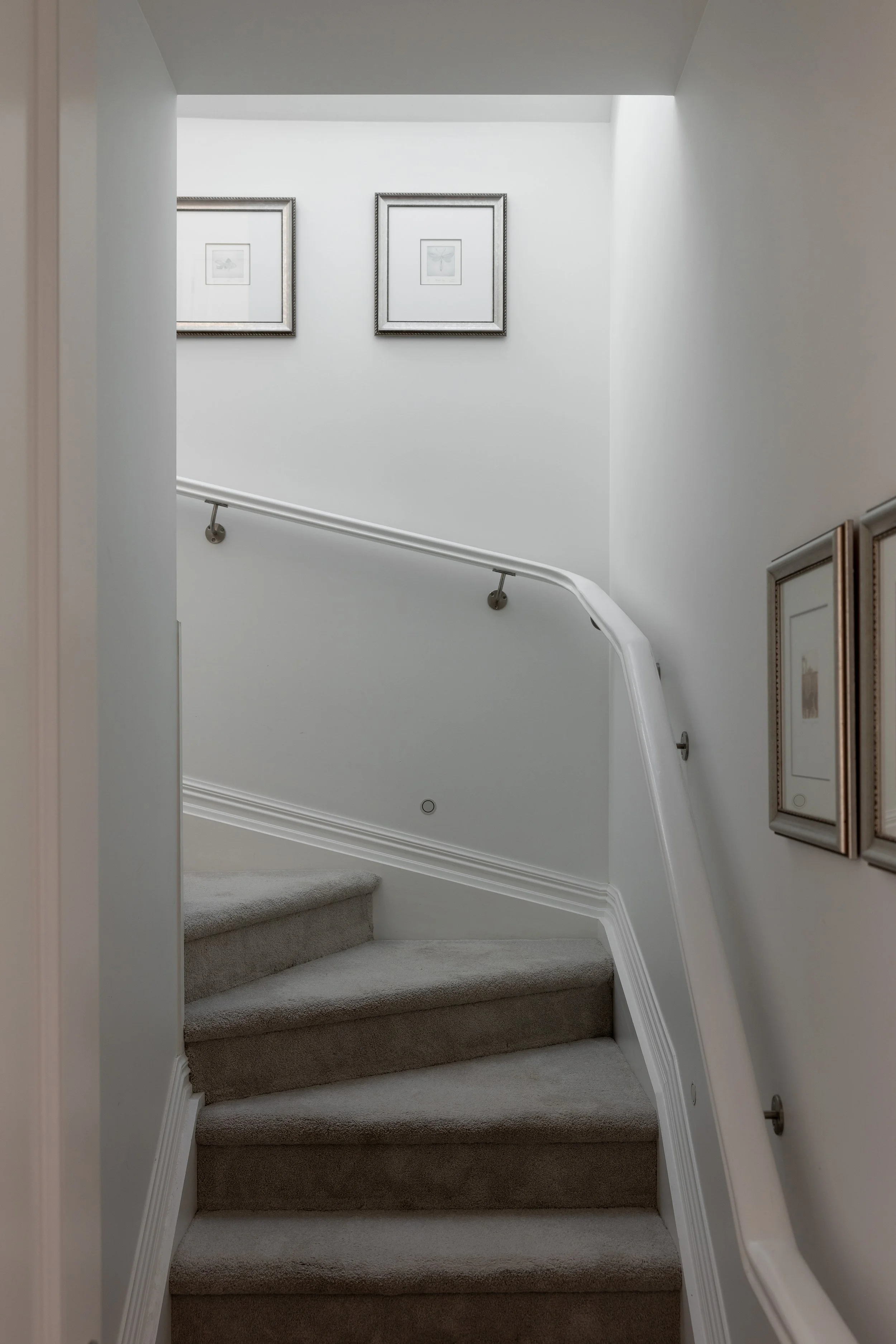 A staircase with carpeted steps and a white curved handrail leading to an upper floor, decorated with framed artwork on the walls, illuminated by natural light from a skylight.