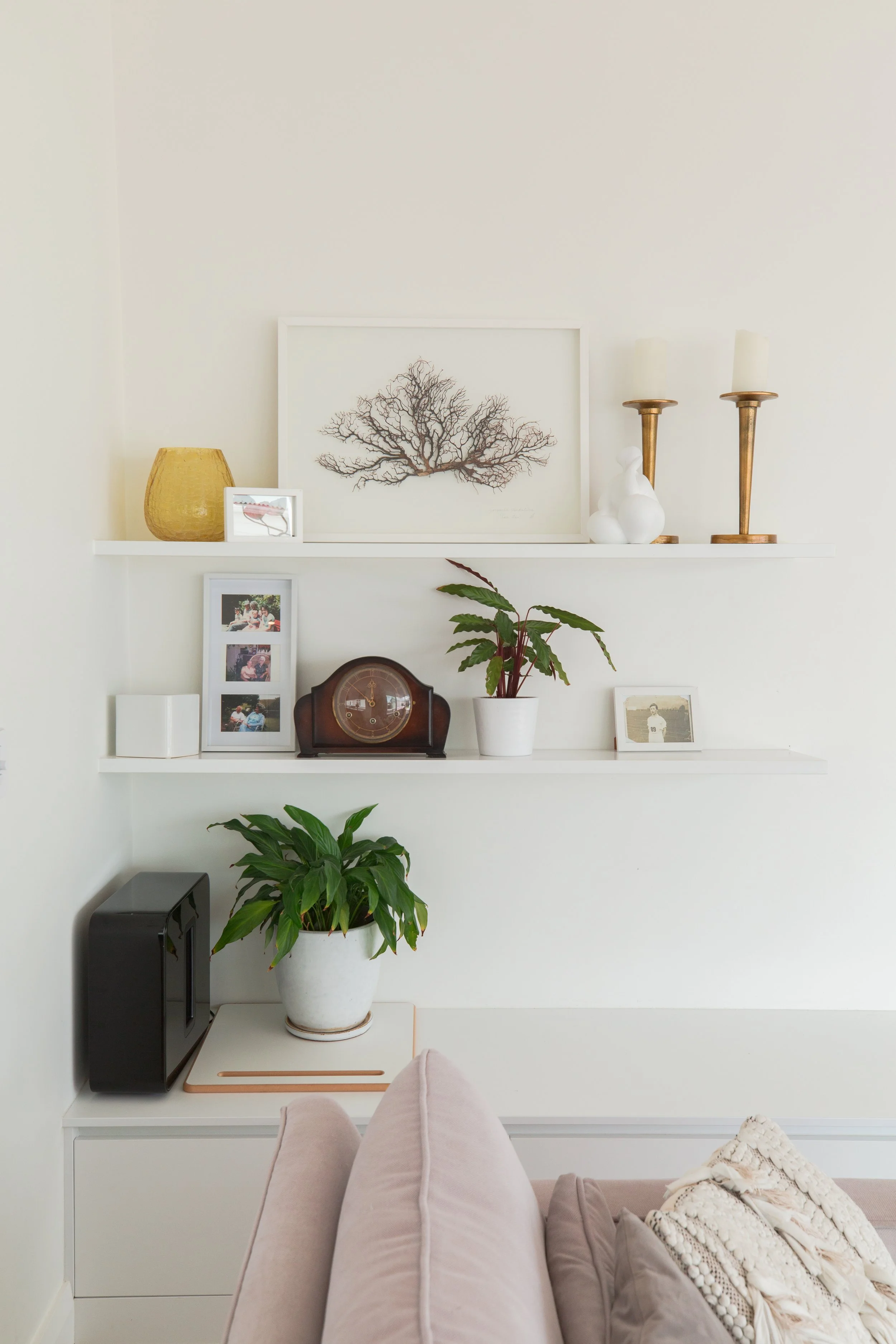 Decorative white wall with two shelves holding framed photos, a potted plant, candles, and decorative objects, next to a beige sofa with throw pillows.