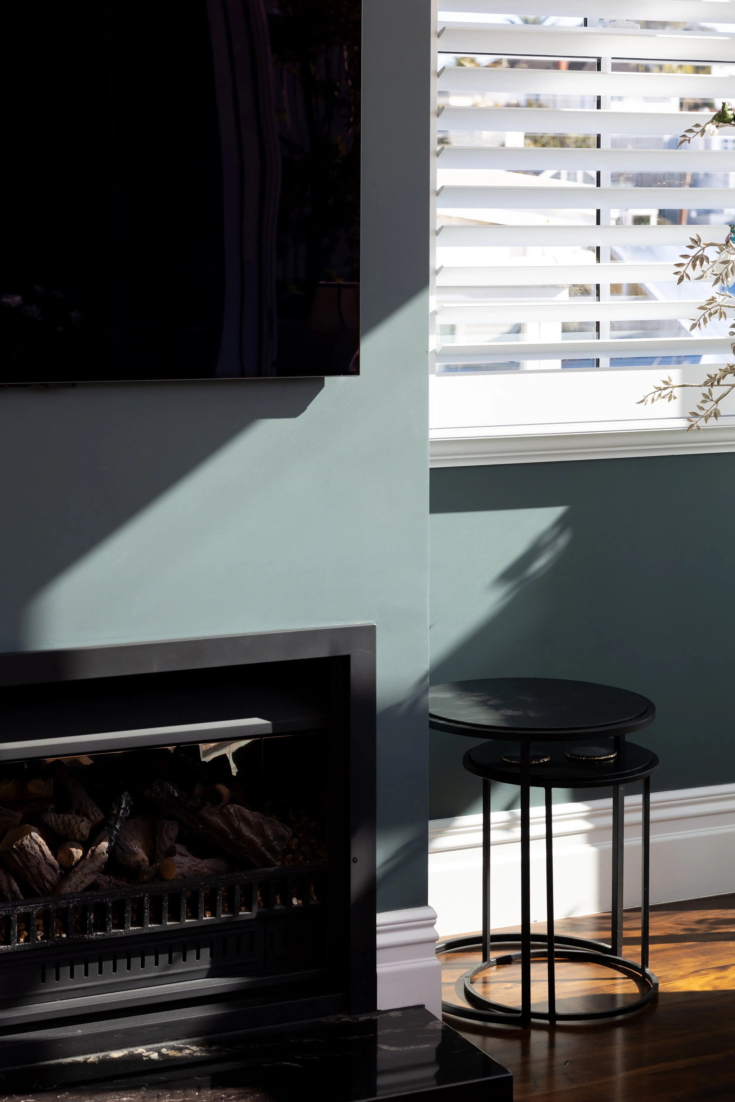 Interior of a living room with a wall-mounted television, a fireplace with logs, and two black nested tables beside a window with white blinds, sunlight casting shadows.