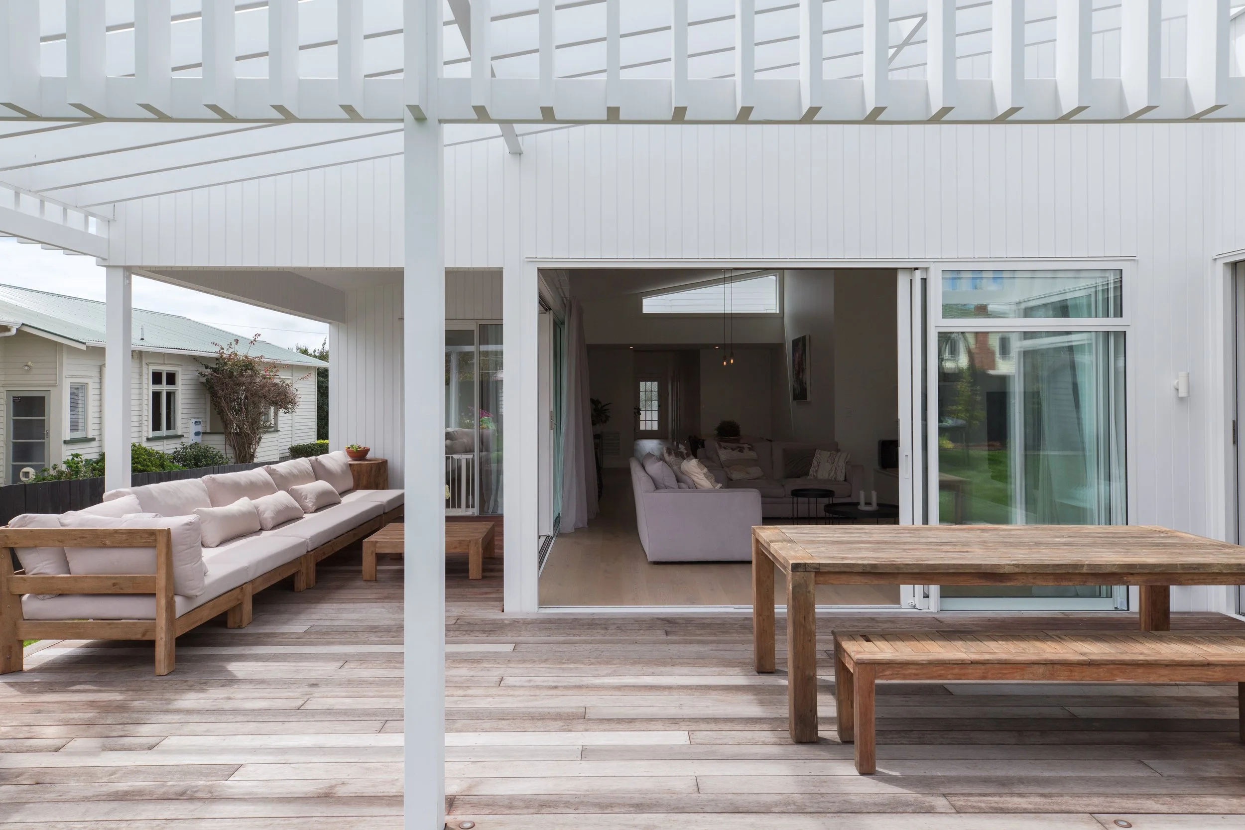 View of an outdoor patio with wooden furniture, including a long cushioned bench and a dining table with benches, connected to an indoor living room through sliding glass doors, under a white pergola.