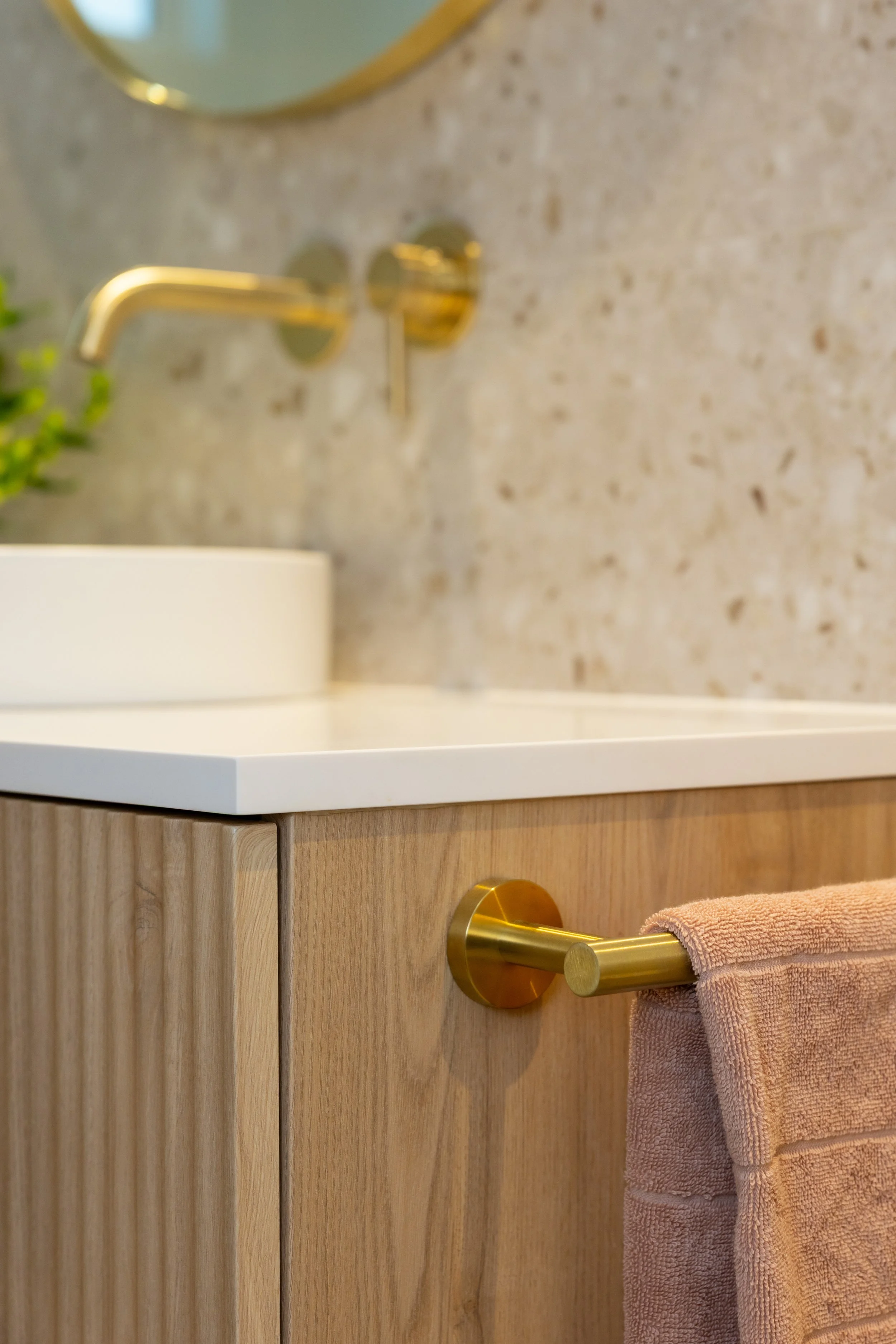 Close-up of a bathroom vanity with a wooden cabinet, a pink towel hanging on a brass towel holder, and a granite countertop. A mirror and brass faucet are visible in the background.