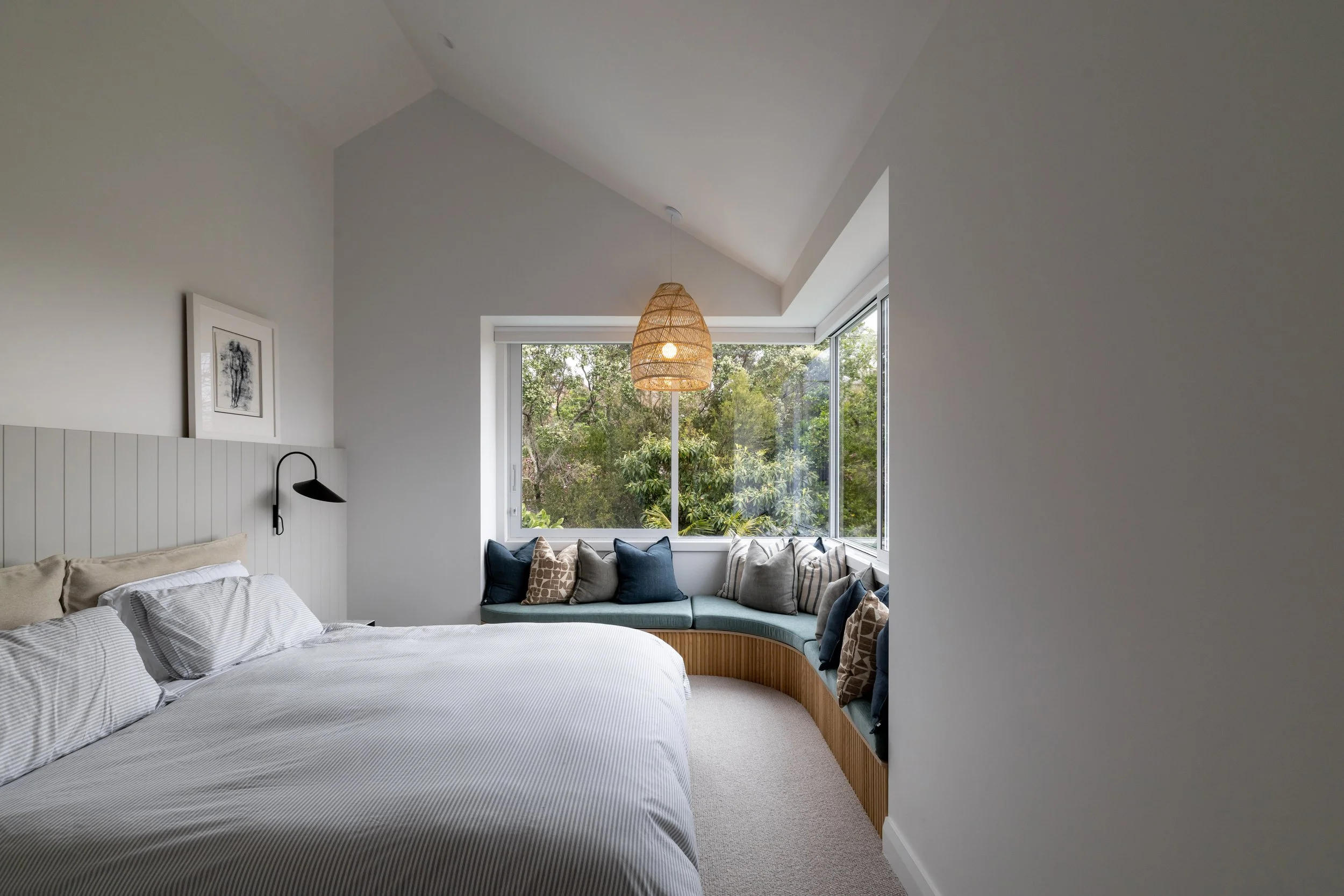 A cozy bedroom featuring a bed with white and gray striped bedding and pillows, a black wall-mounted reading lamp, a surrounded window with a view of green trees, a wicker hanging light fixture, and a built-in window seat with various blue, gray, and patterned throw pillows.