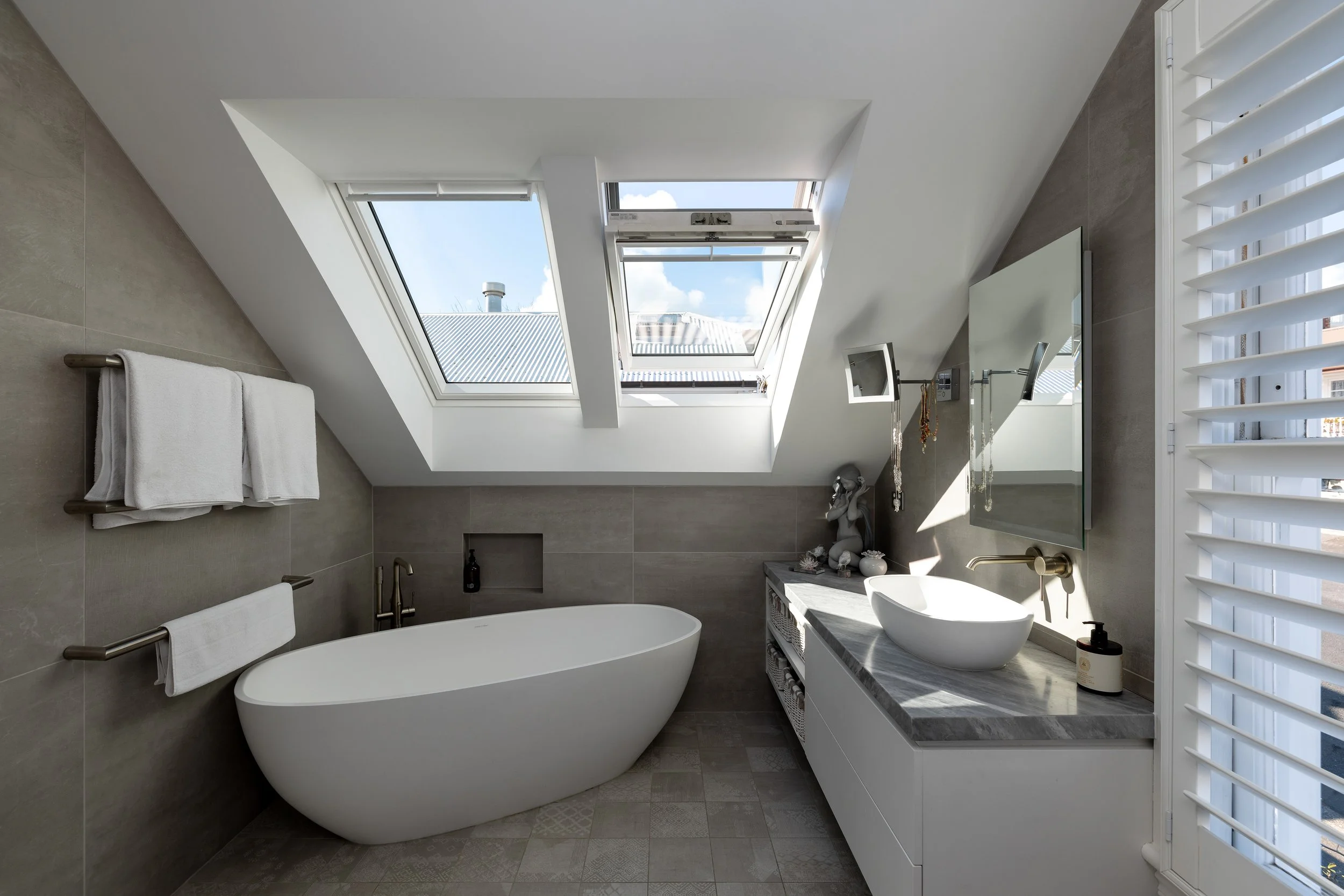 Modern bathroom with bathtub under skylight, gray tiled walls, and a vanity with a vessel sink and mirror.