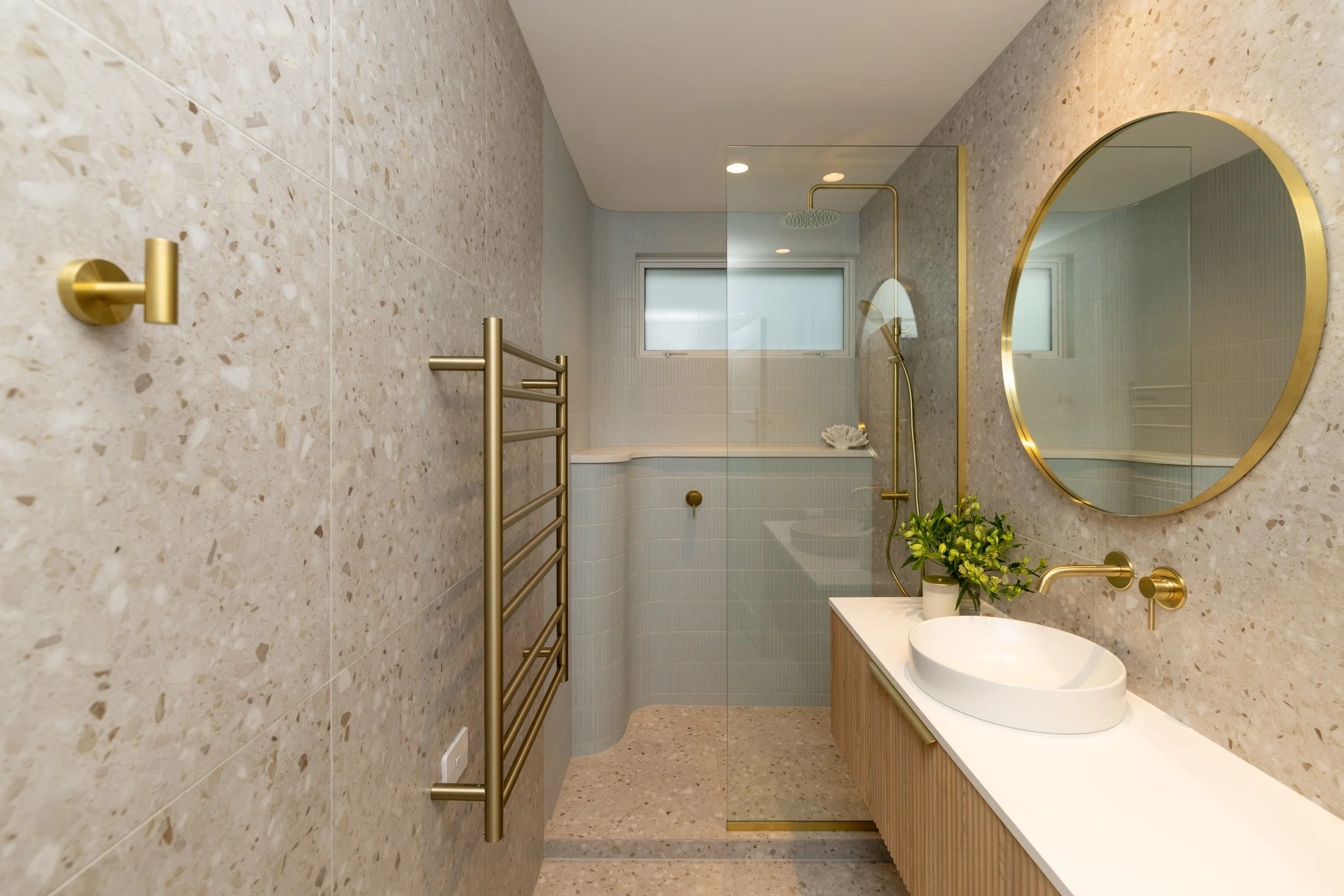 Modern bathroom with terrazzo walls, a round mirror with a gold frame, a vessel sink on a light wood vanity, potted plant, brass fixtures, shower with a glass partition, and a small window.