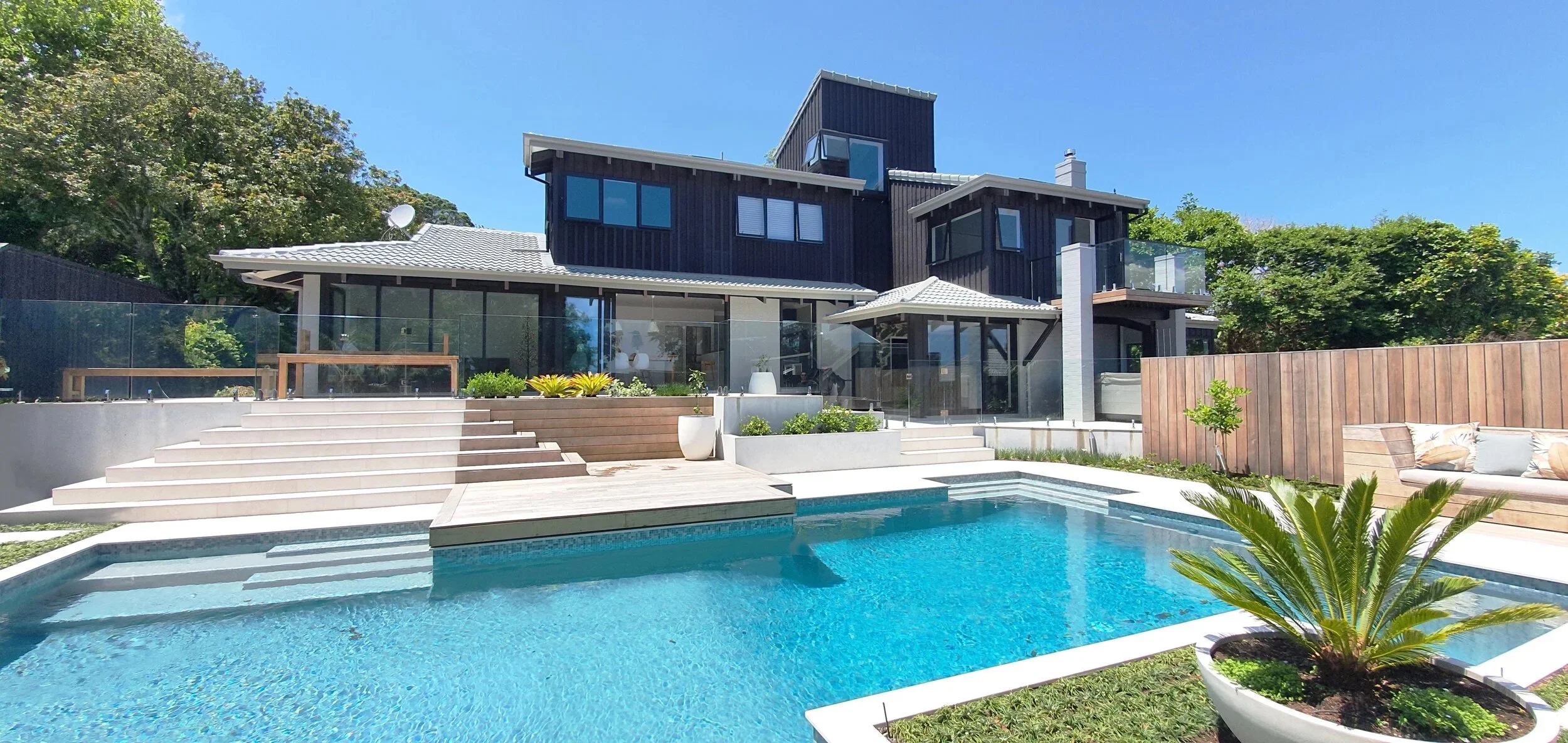Modern multi-level house with black siding, large glass windows, and a white tiled roof. In the foreground, there's a swimming pool with steps leading into it, surrounded by a patio with seating and potted plants. The backyard features a landscaped garden with a wooden privacy fence and trees in the background under a clear blue sky.