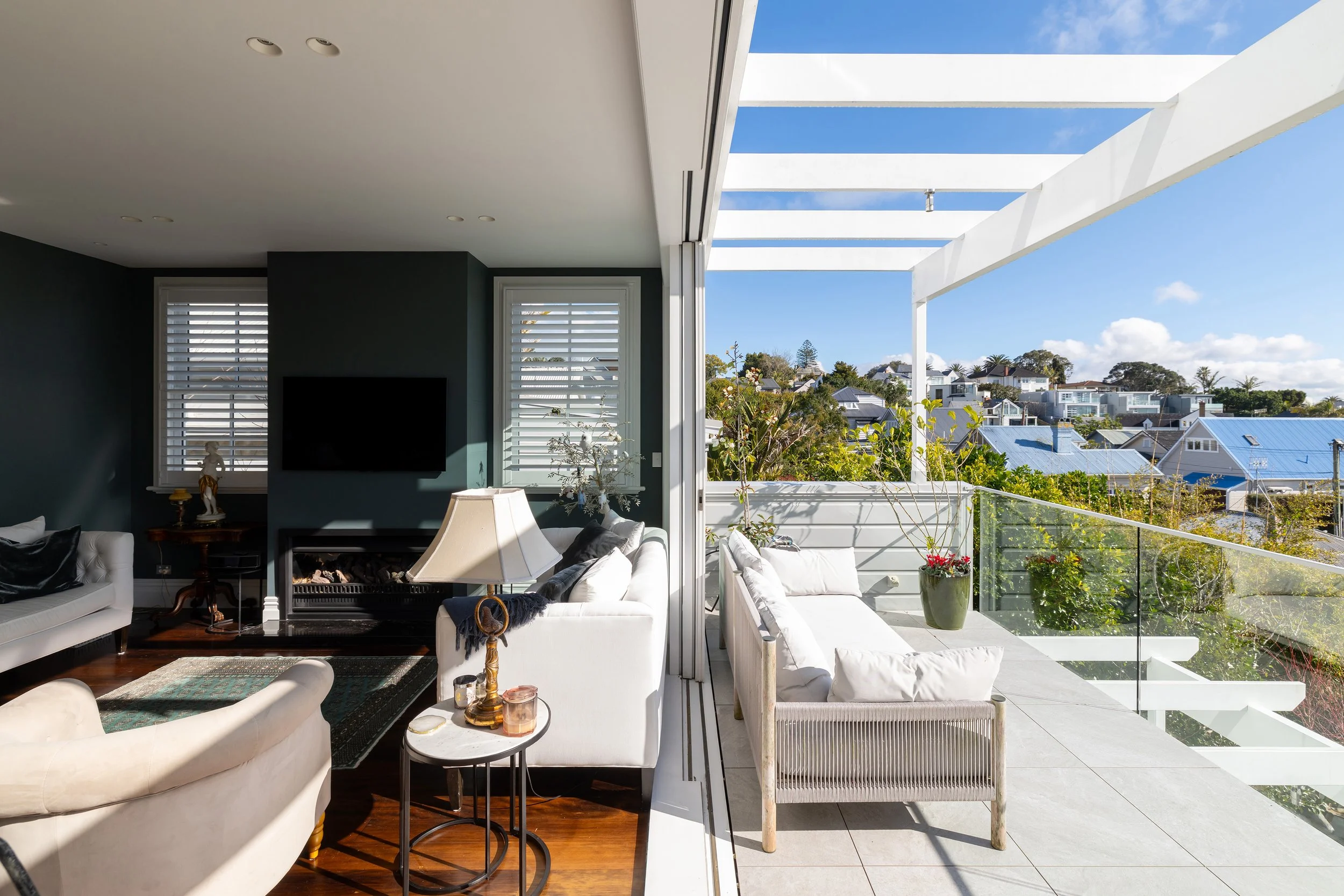 Interior living room with dark green walls, white sofas, a lamp, and a fireplace. Adjacent to the living room is an outdoor balcony with white furniture, potted plants, glass railing, and a view of houses and trees under a blue sky.