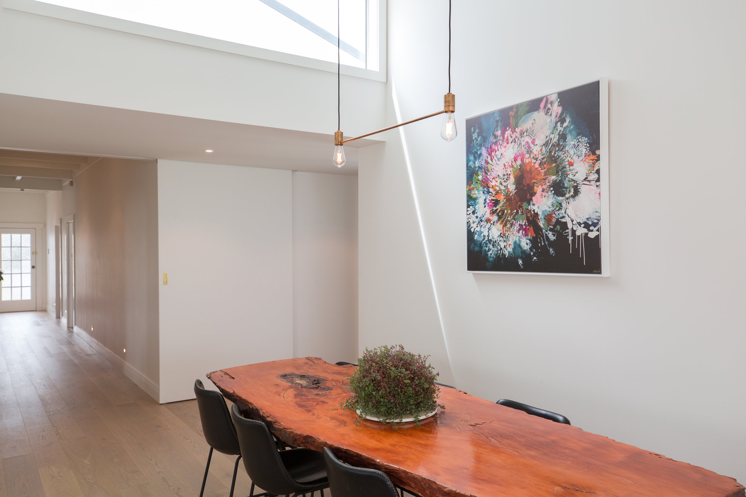 Modern dining room with a large live edge wooden table, black chairs, abstract floral artwork on white wall, and hanging light bulbs, natural light from a nearby window.