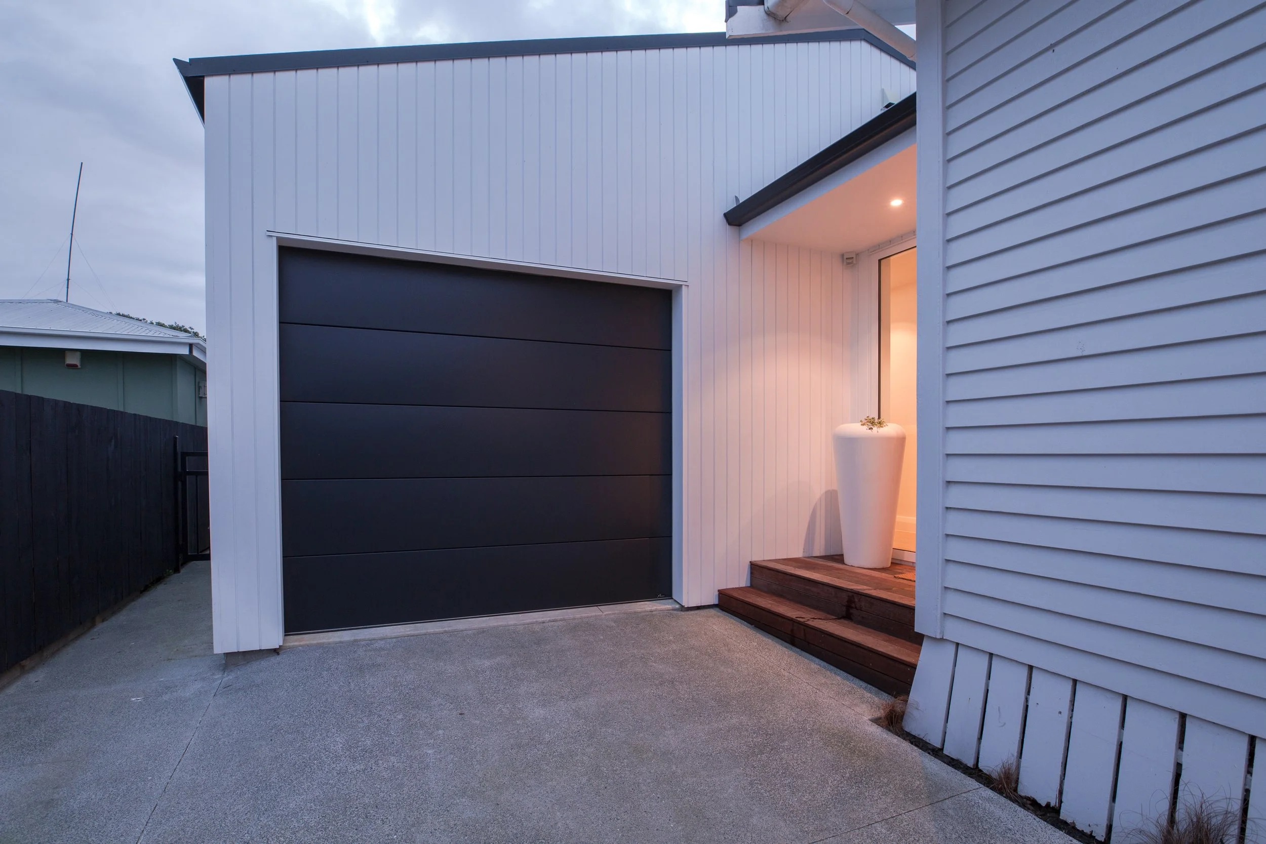 Exterior view of a modern house with a black garage door, wooden steps, and a large white planter with a plant near the entrance, during evening time.
