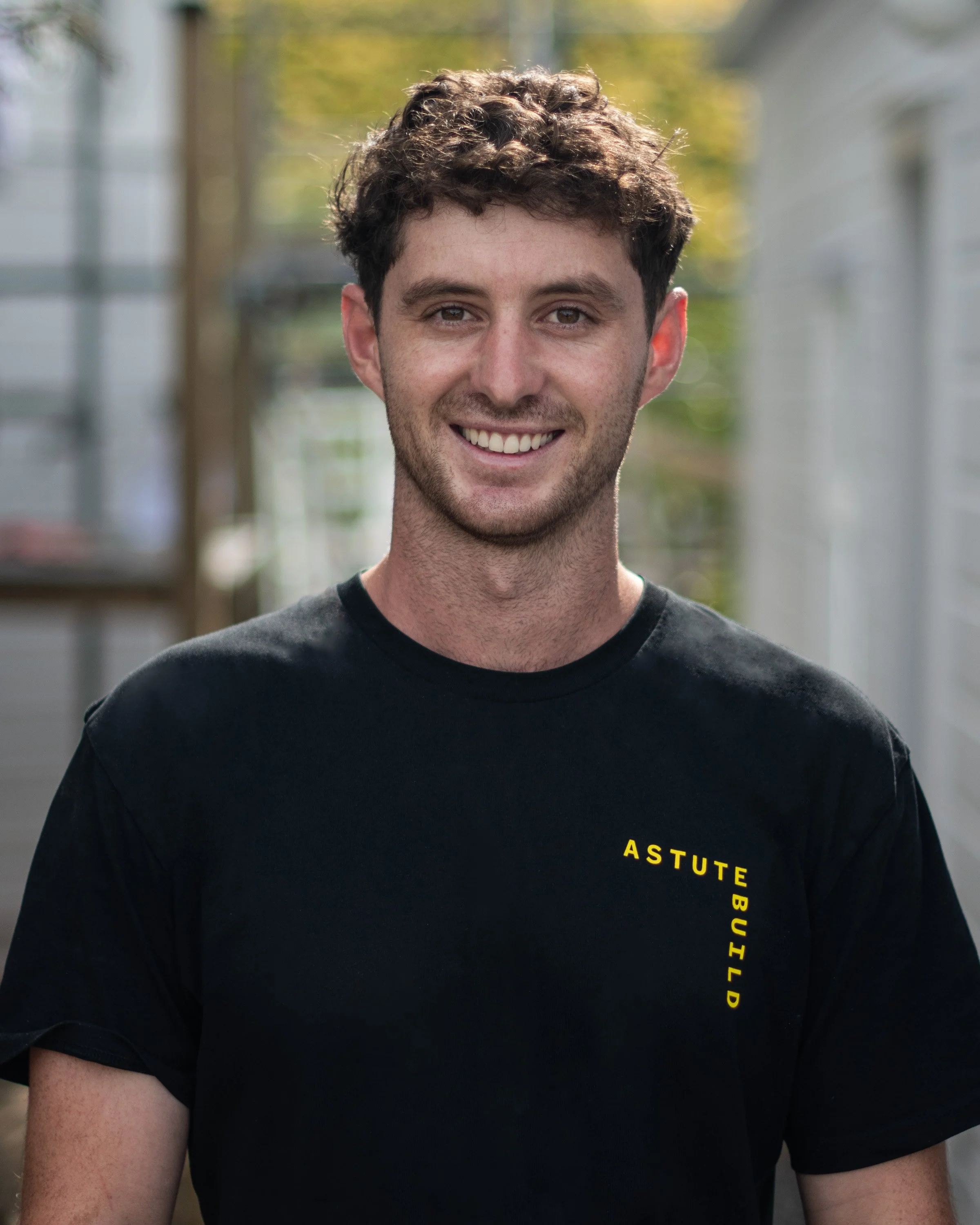 A smiling young man with curly brown hair, wearing a black T-shirt with yellow text reading "ASTUTE BUILD" on the chest, standing outdoors with a blurred background of trees and buildings.