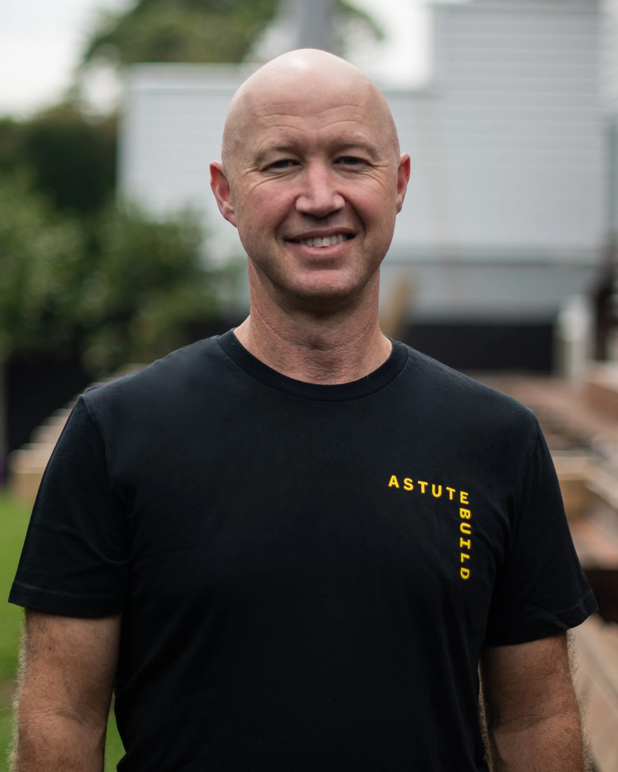 A smiling bald man wearing a black t-shirt with yellow text that reads 'ASTUTE BUILD' standing outdoors with a blurred background of a house and greenery.