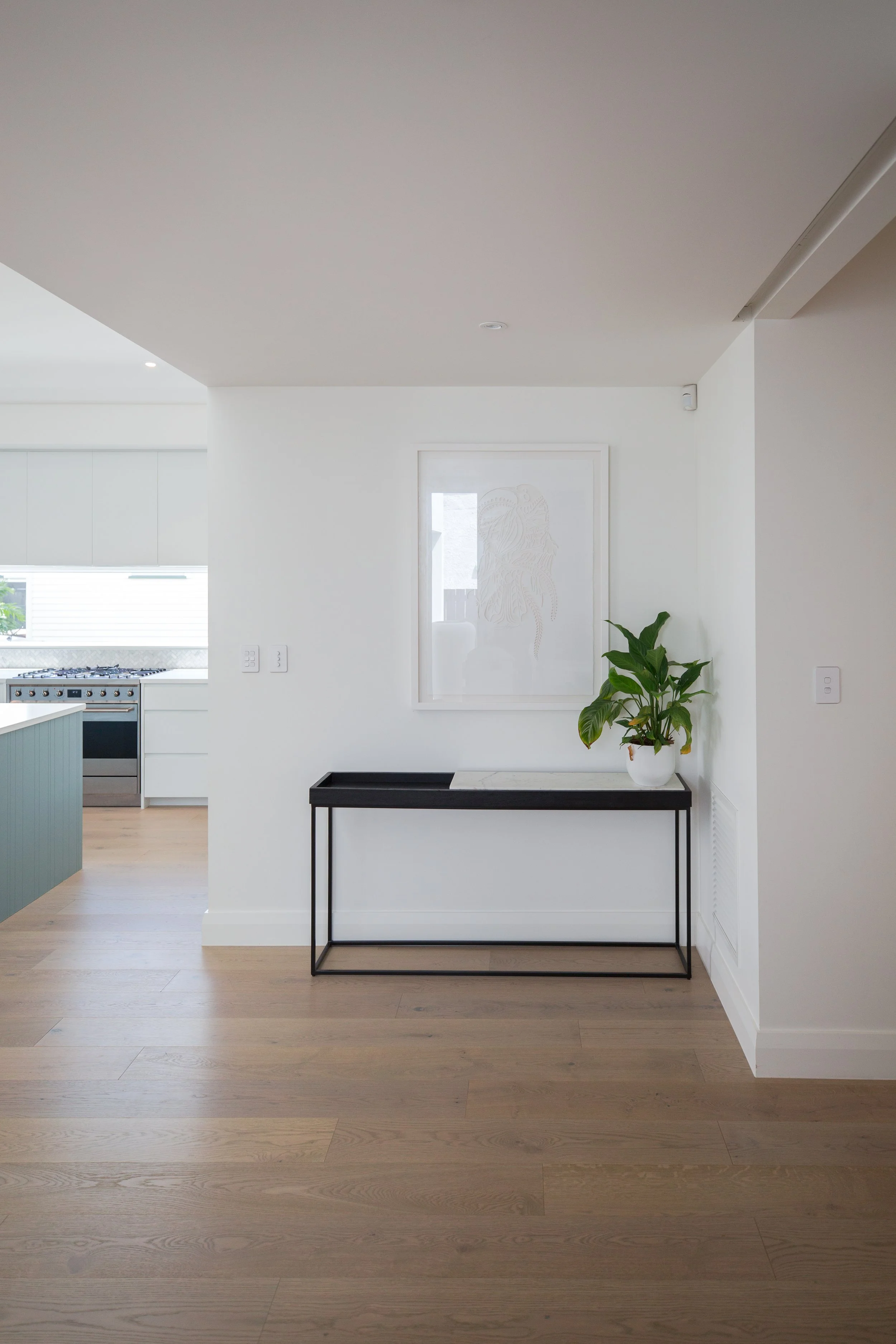 Minimalist interior with a black console table and a potted plant near a white wall, with a framed line drawing and a kitchen area in the background.