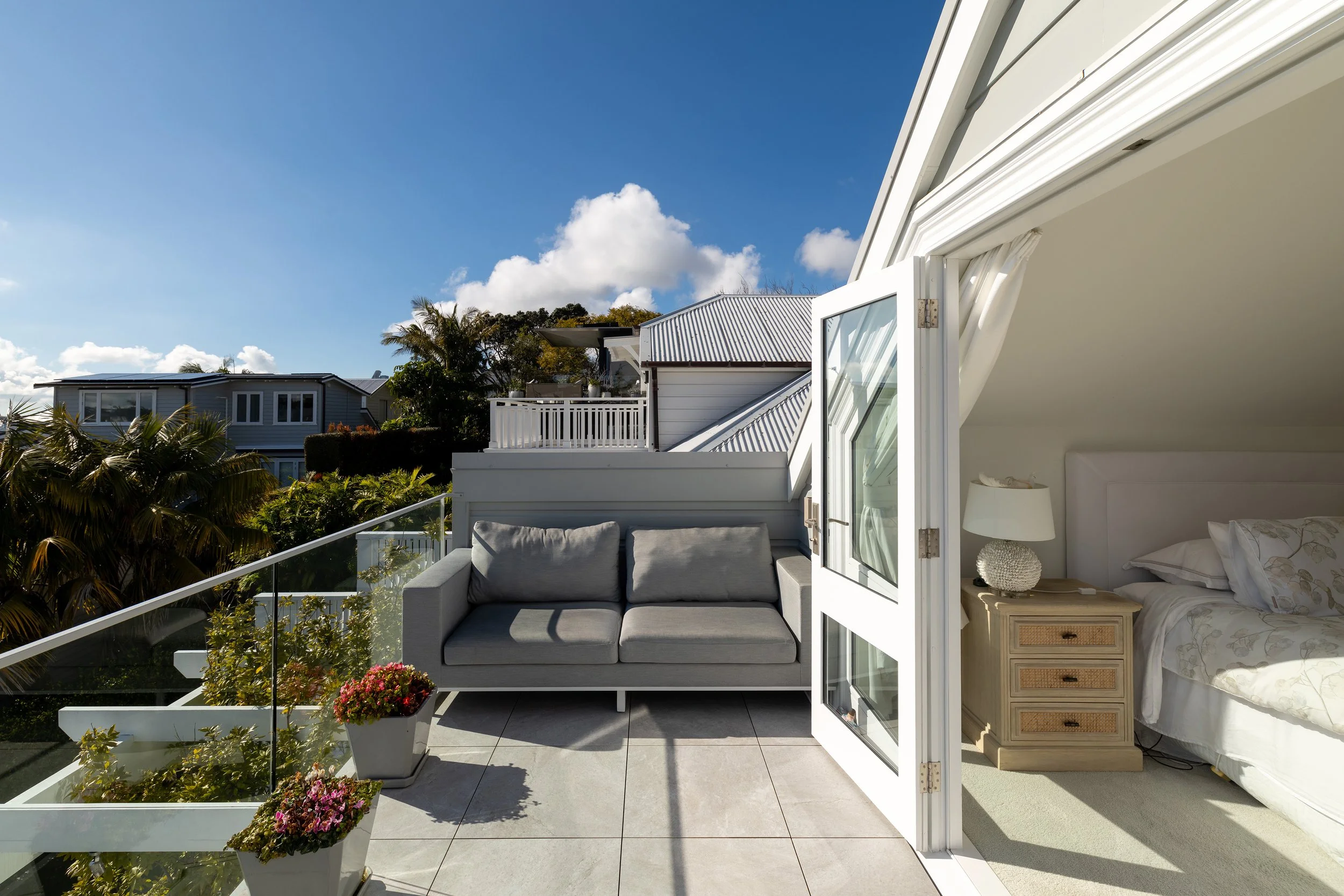 Balcony view of a residential neighborhood with a gray outdoor sofa and potted flowers, overlooking houses with white roofs on a sunny day.