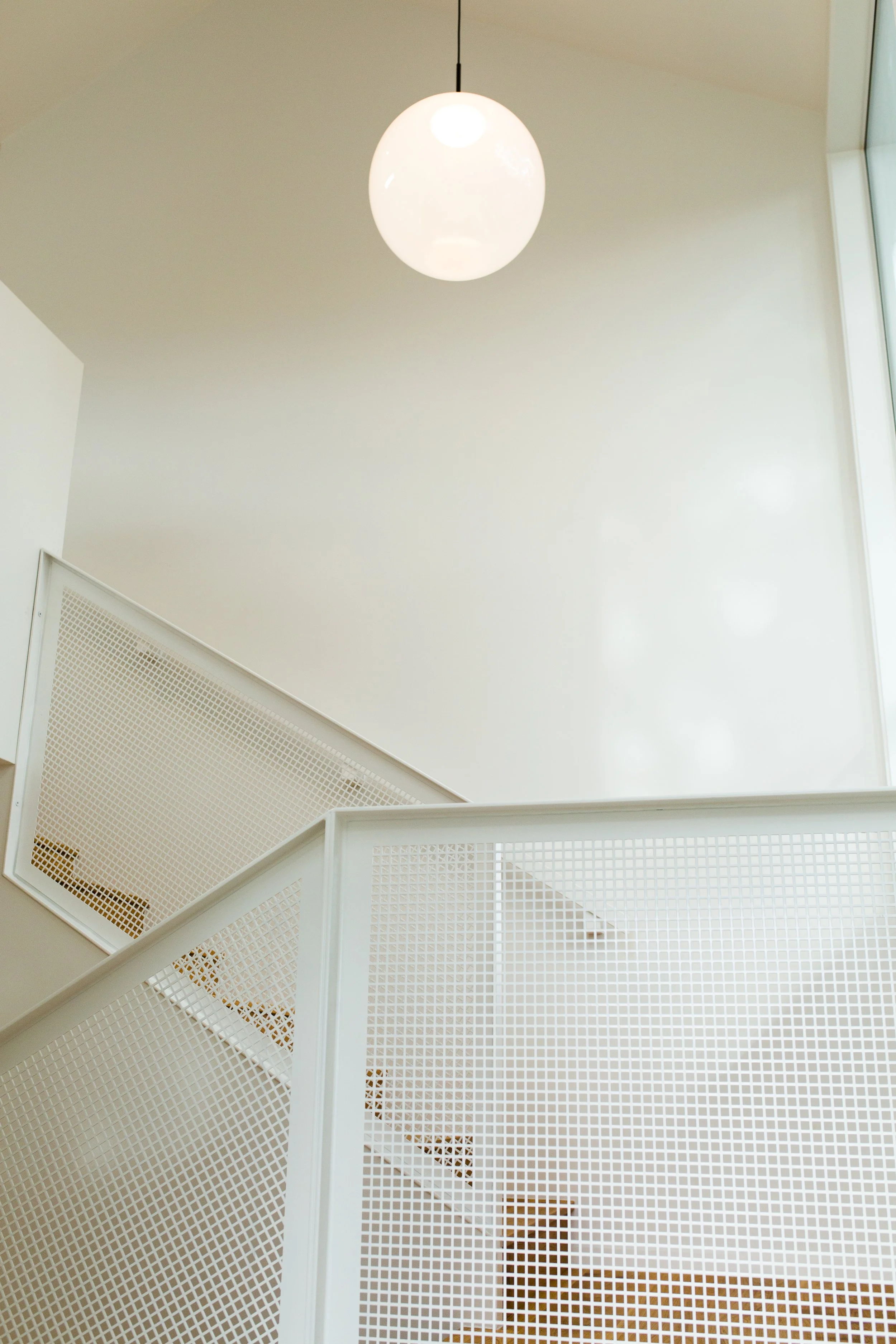 Interior shot of a staircase with white metal railings and a large round white hanging light fixture, near a corner with a large window.