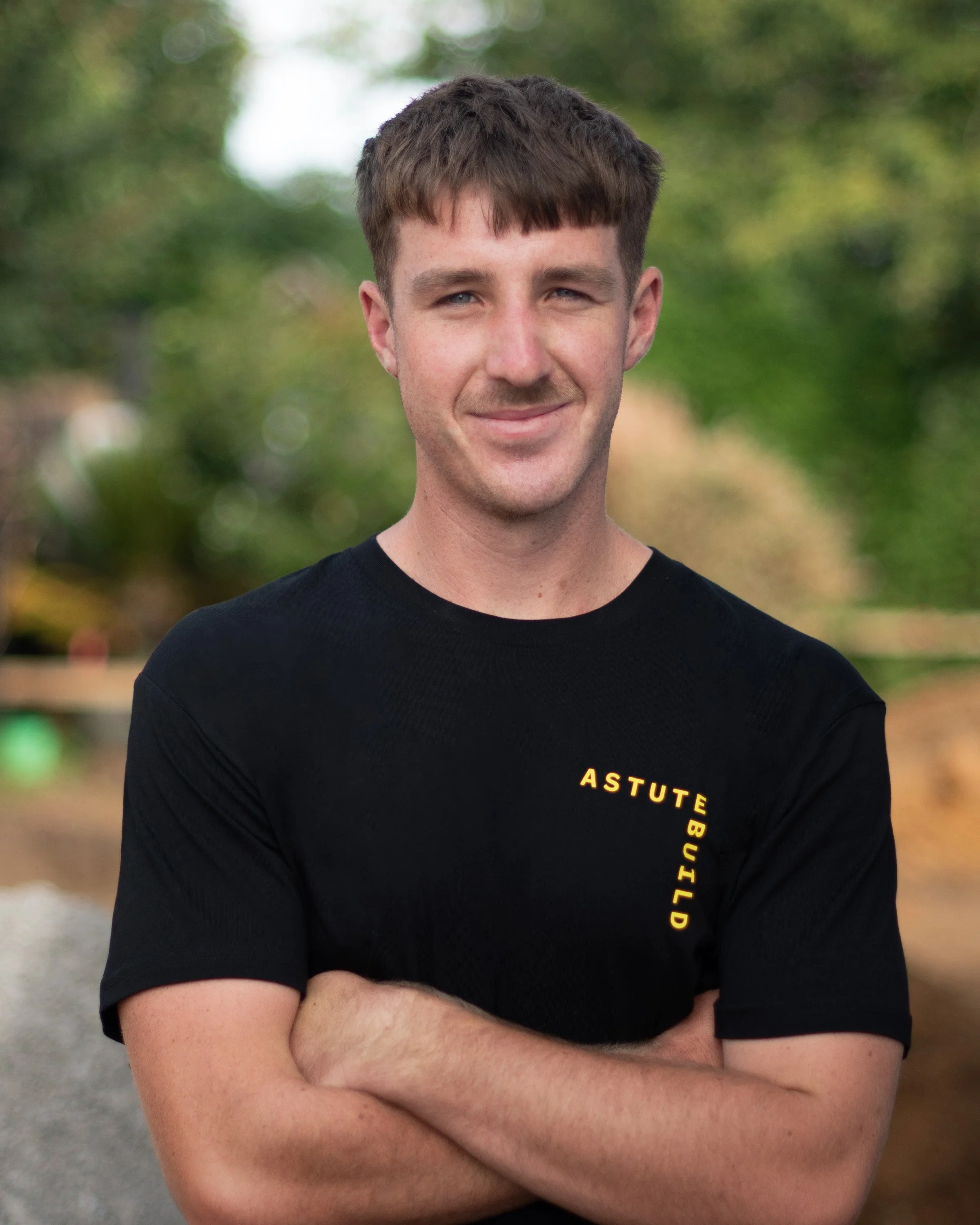 A young man with short brown hair, wearing a black T-shirt with yellow text reading 'ASTUTE BUILD', standing outdoors with trees and blurred background.