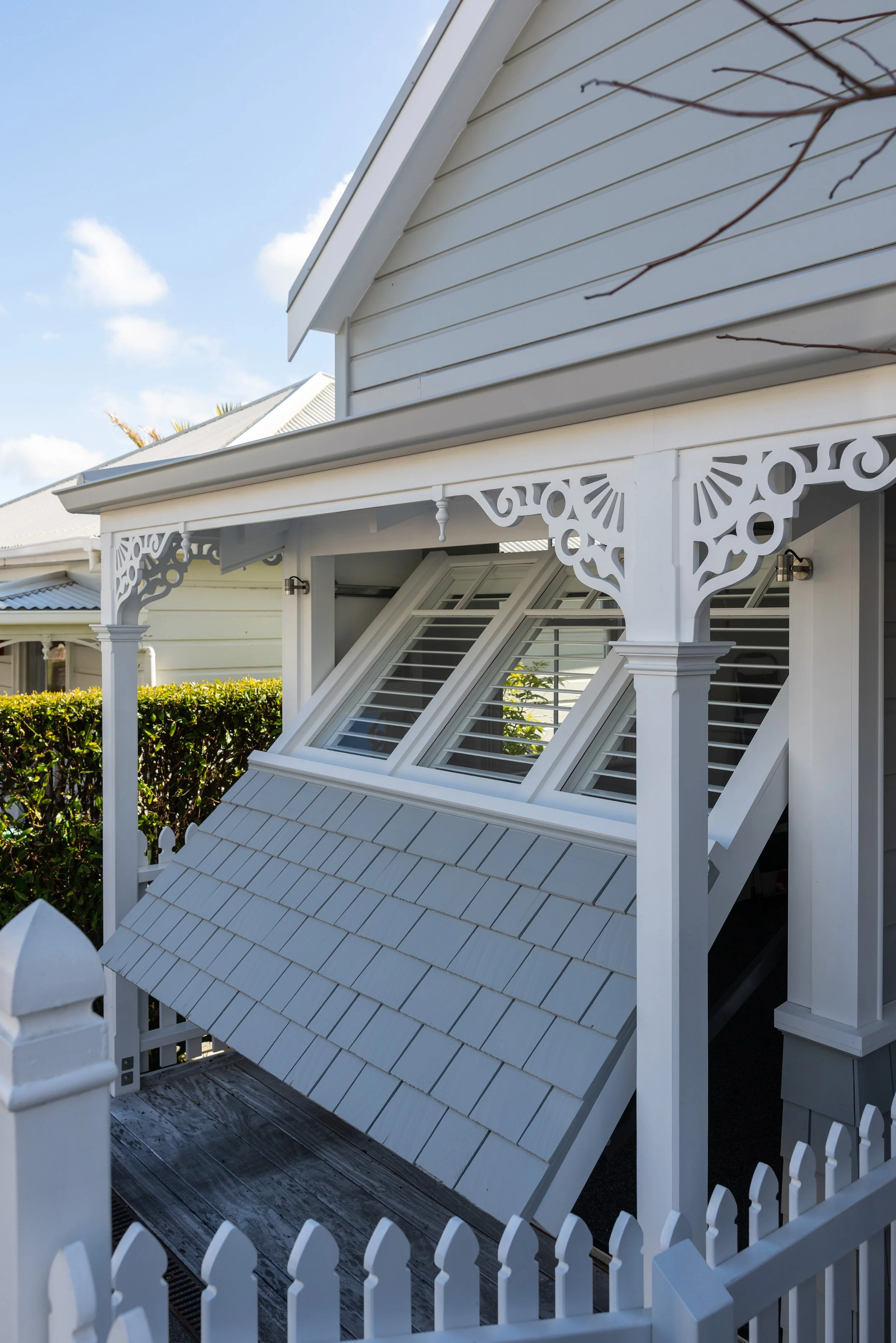 A white house with an ornate porch and decorative trim, featuring attic windows with angled shutters, a small fenced patio, and a hedge in the background under a partly cloudy sky.