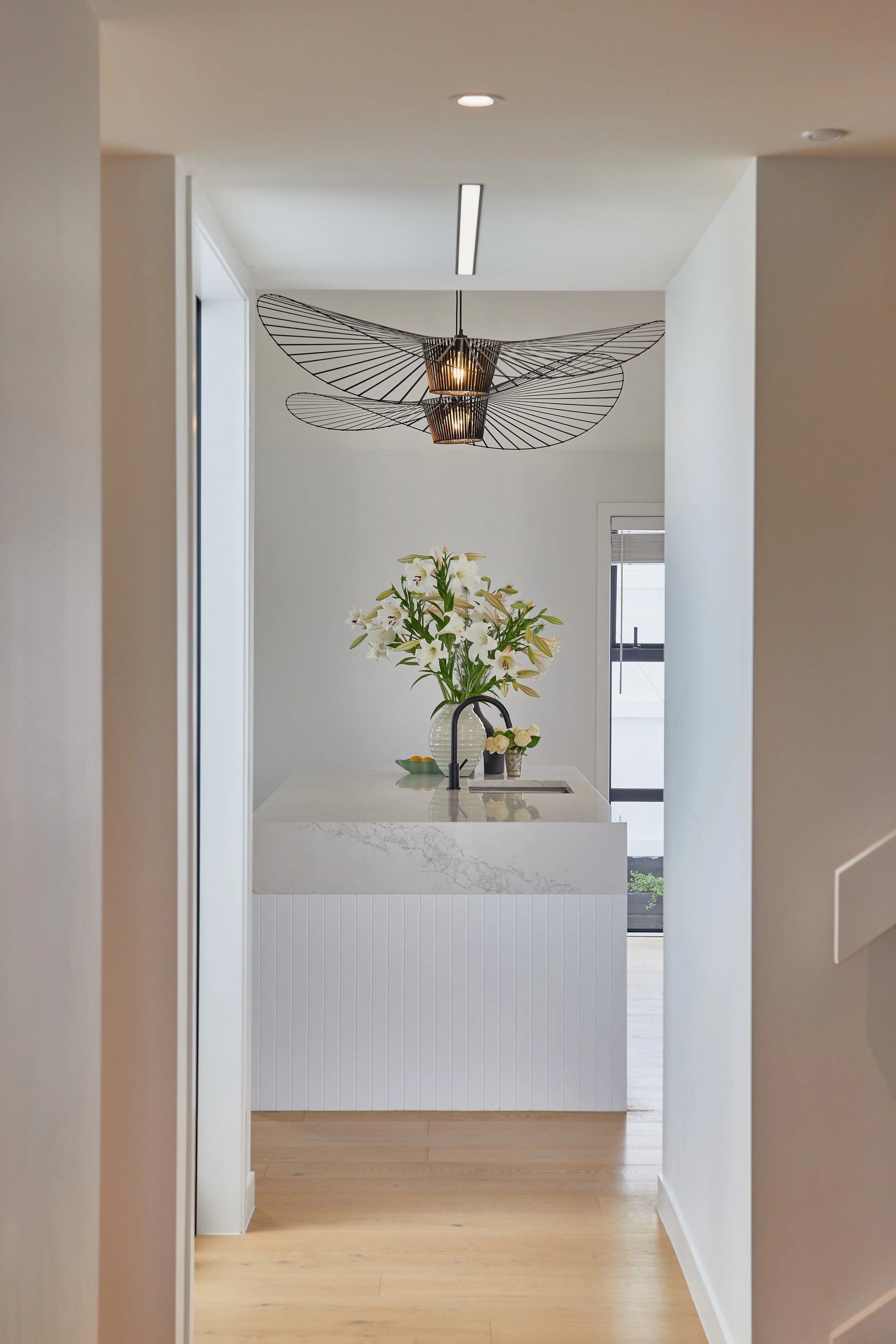 Modern kitchen with white island, vase of white lilies, black faucet, and an artistic black wire chandelier hanging from the ceiling.