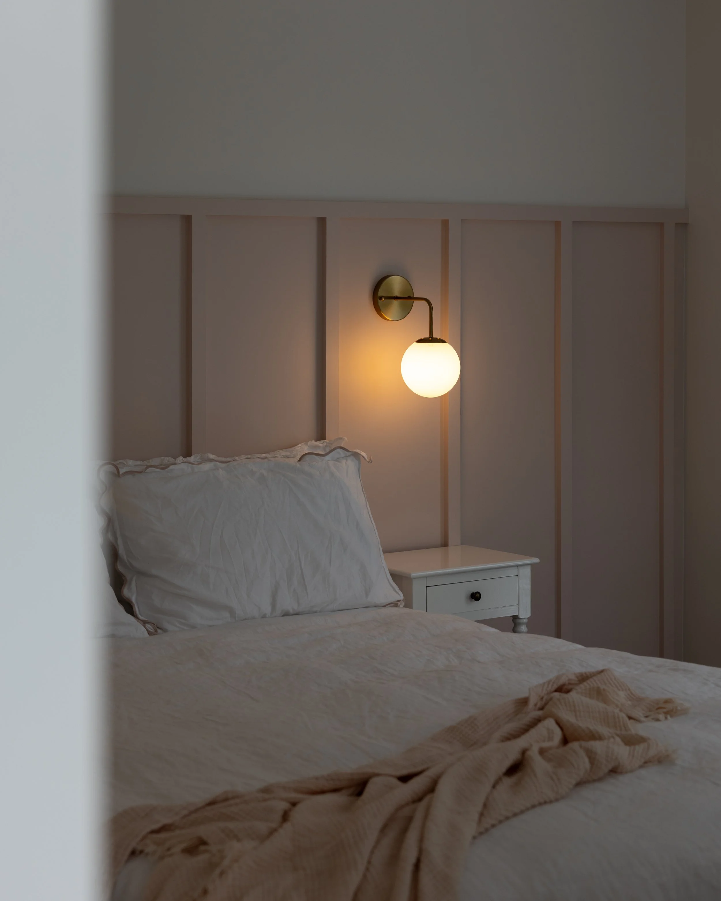 A bedroom scene with a bed and a white pillow, a nightstand, and a wall-mounted light fixture with a glowing round bulb.