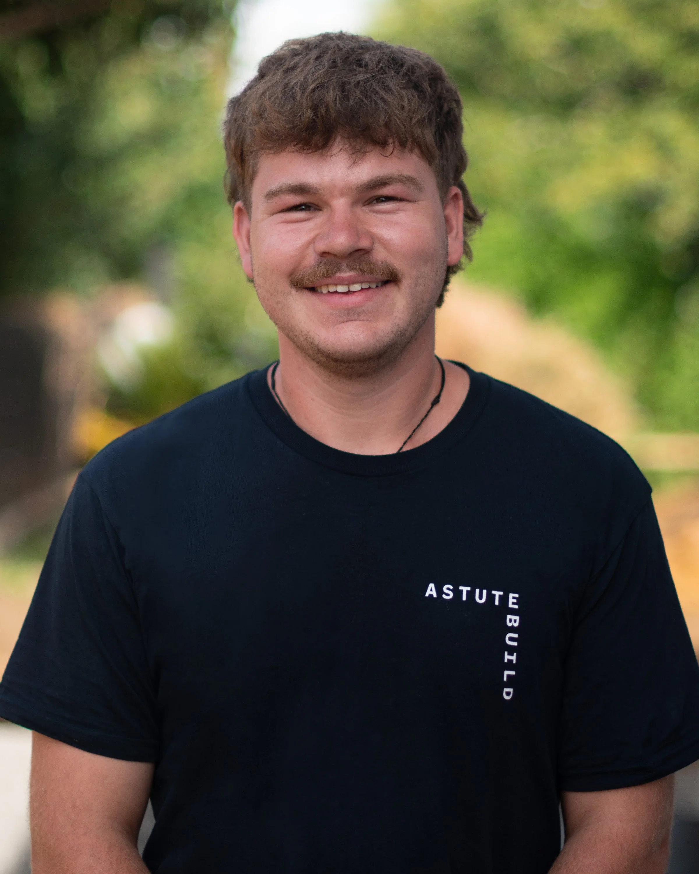 A young man with curly brown hair, a mustache, and a beard, smiling outdoors with a blurred green and yellow background, wearing a black T-shirt that says 'ASTUTE BUILD'.