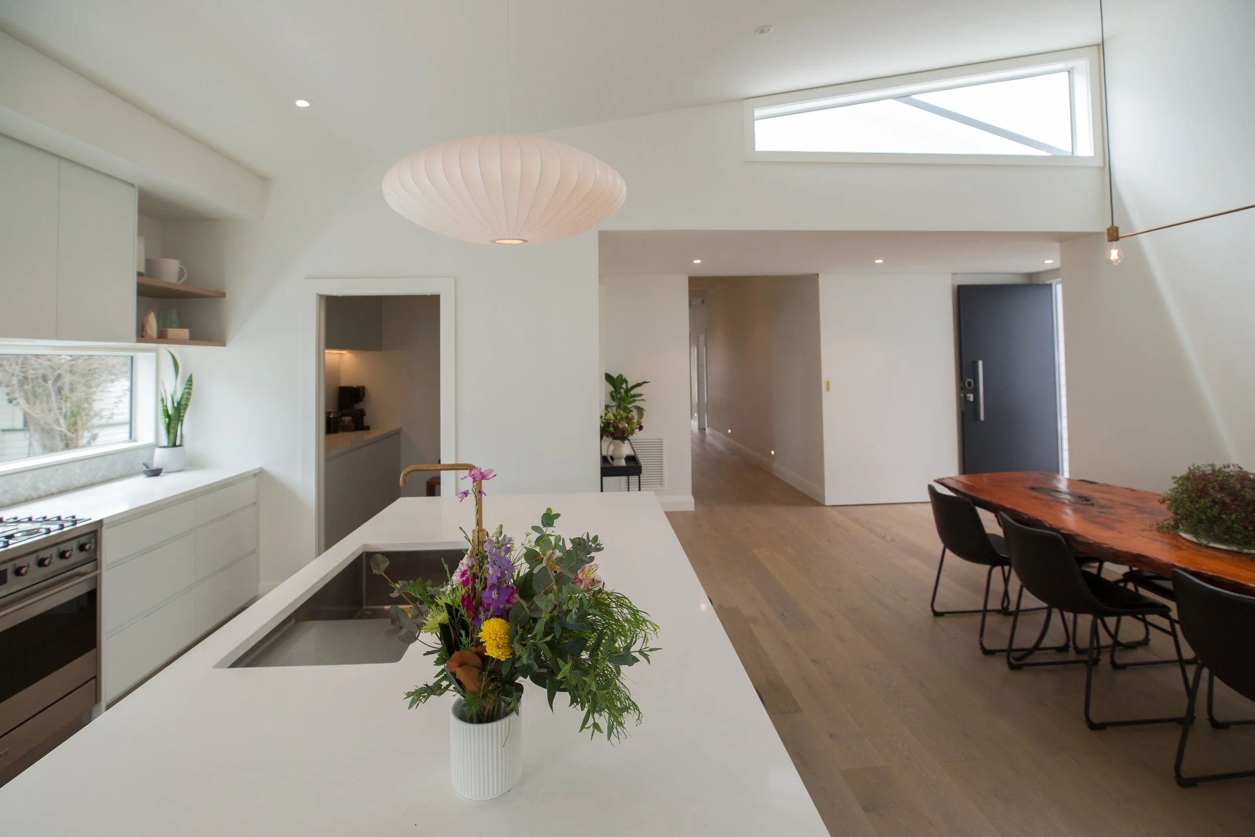 Modern open-concept kitchen and dining area with white cabinets, a white island with a flower arrangement, a wooden dining table with black chairs, hardwood floors, and large windows with natural light.