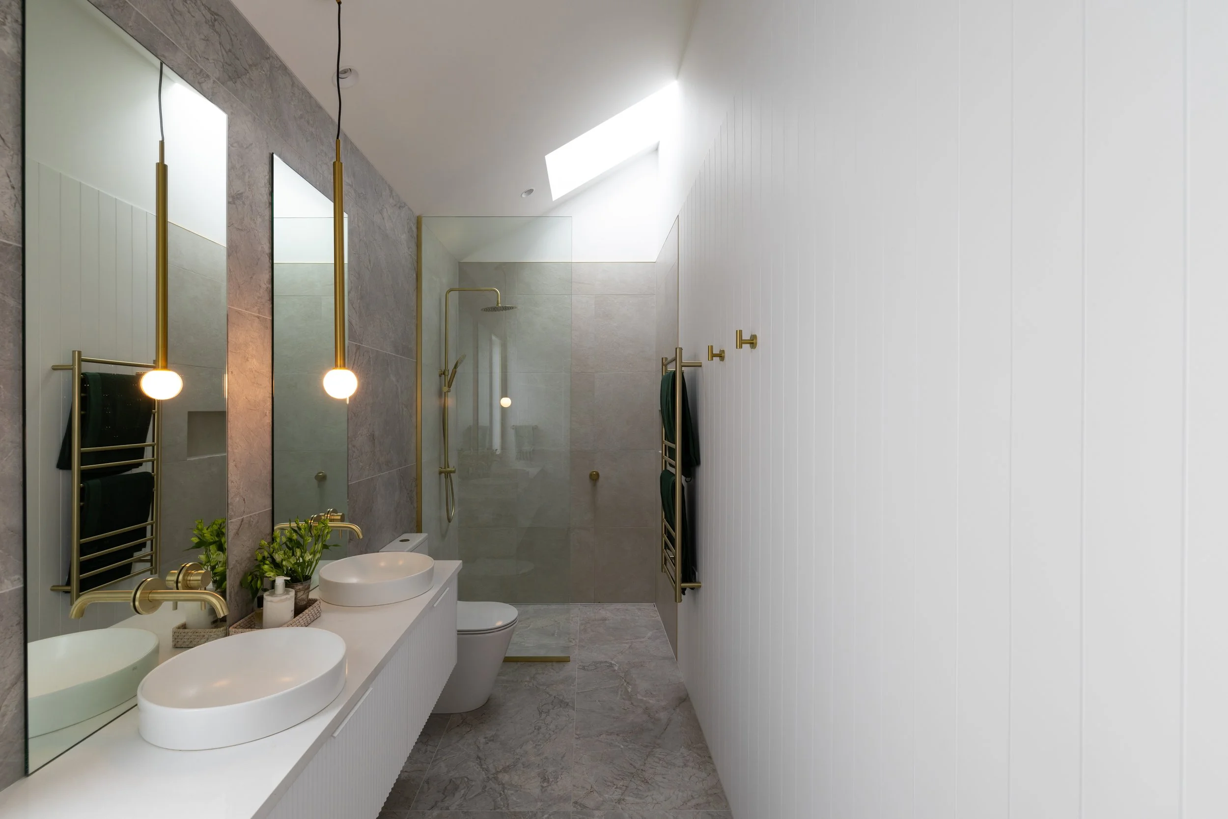Modern bathroom with double white vessel sinks, gold fixtures, gray marble flooring, large mirrors, a glass-enclosed shower with gold fixtures, white paneled wall, and natural light from a skylight.