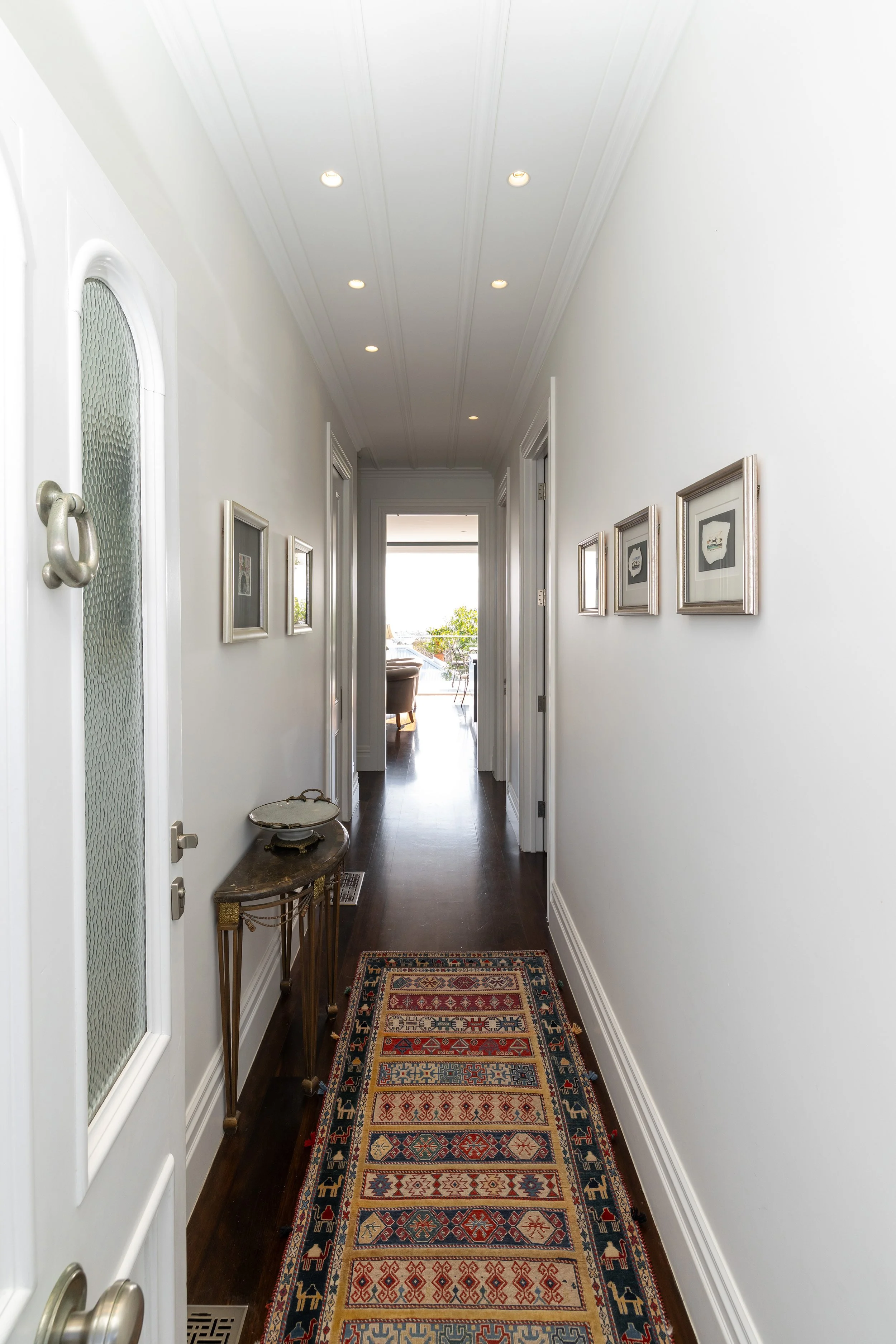 White hallway with framed pictures on the wall, a small antique table on the left, and a colorful patterned runner rug on the floor, leading to a bright room with view outside.