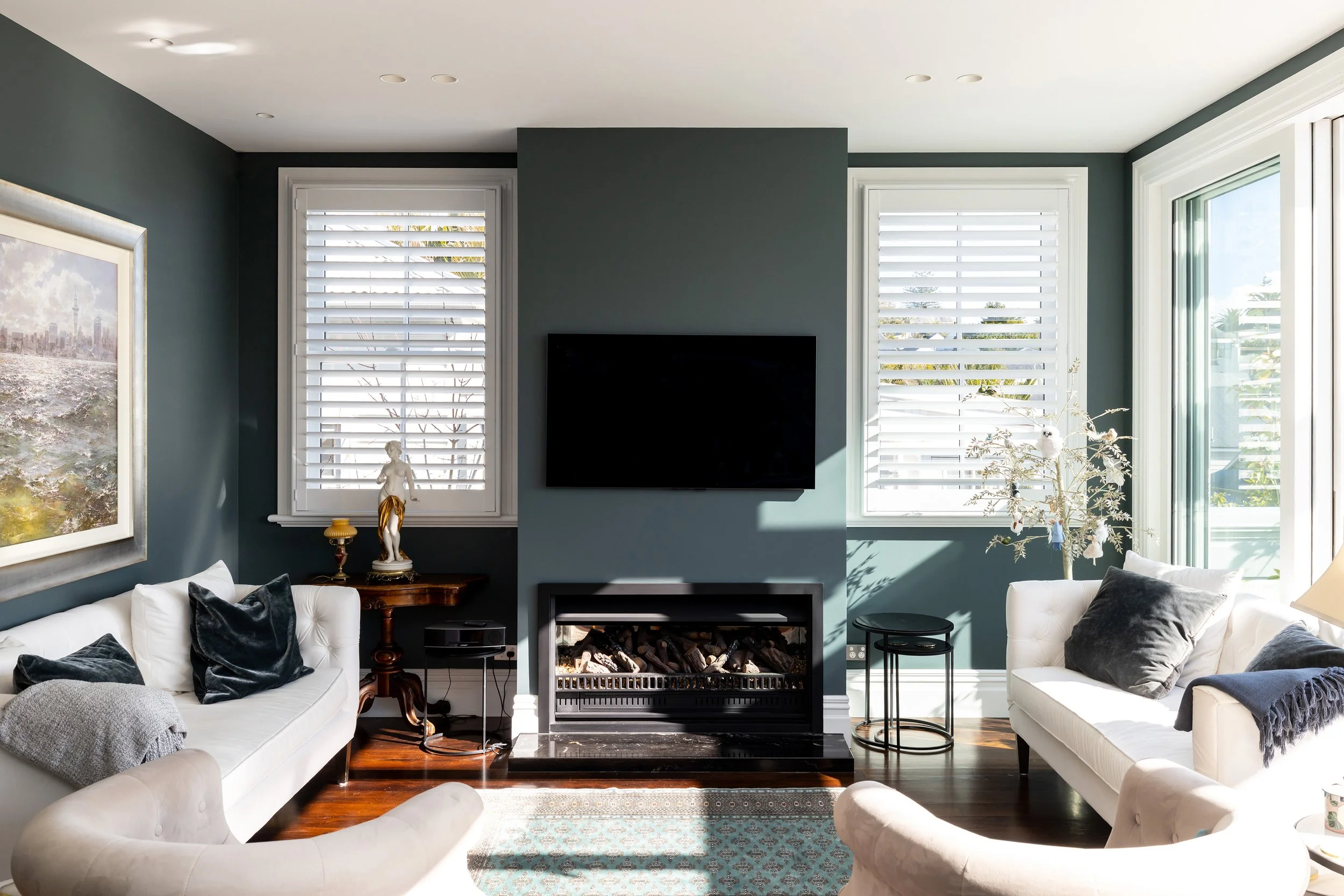Living room with white sofas, dark hardwood flooring, blue walls, two large windows with white blinds, a wall-mounted TV above a fireplace, and a sliding glass door to the right, letting in natural light.