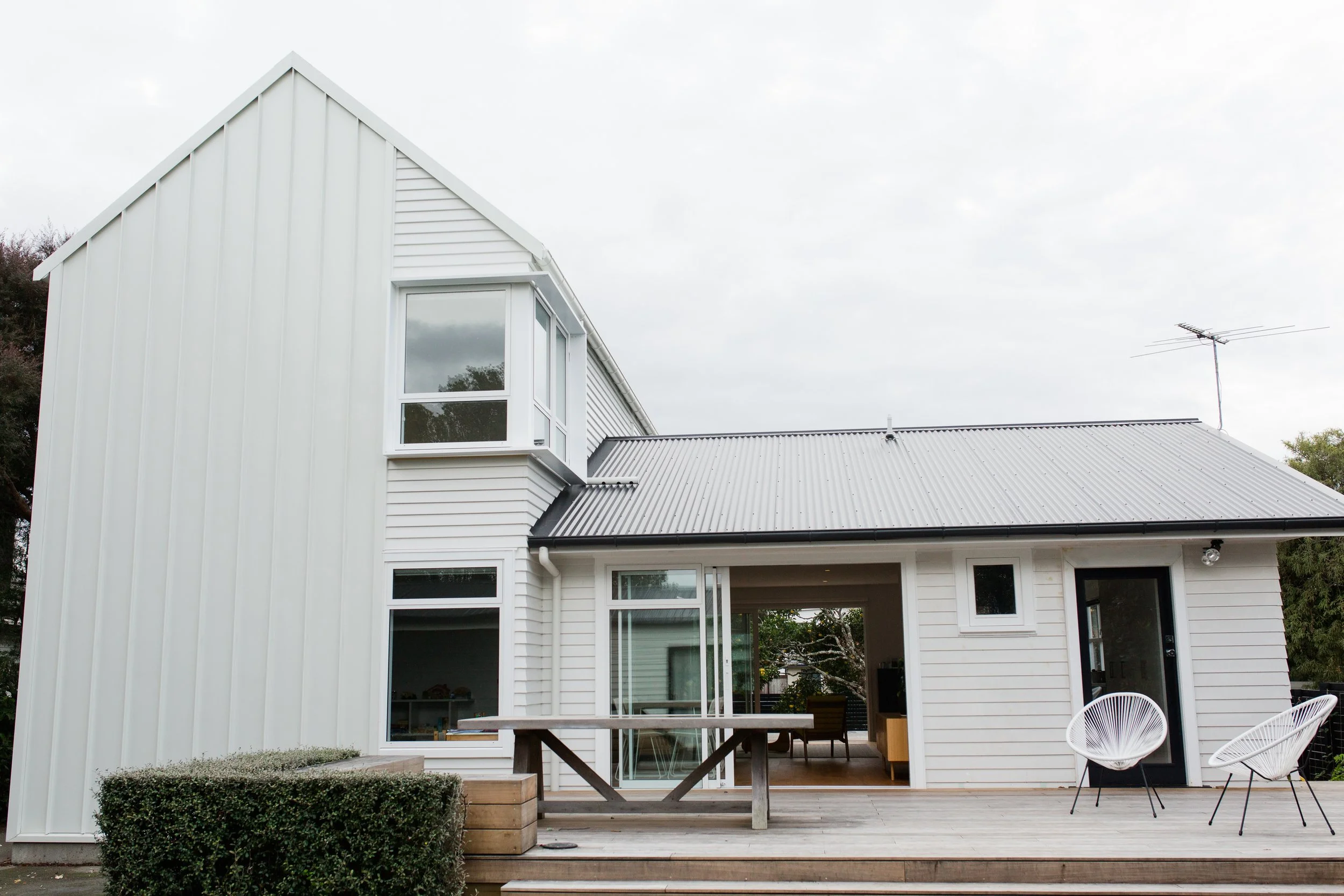 Modern two-story house made of white siding with a metal roof, large windows, and an outdoor deck with chairs and a table.