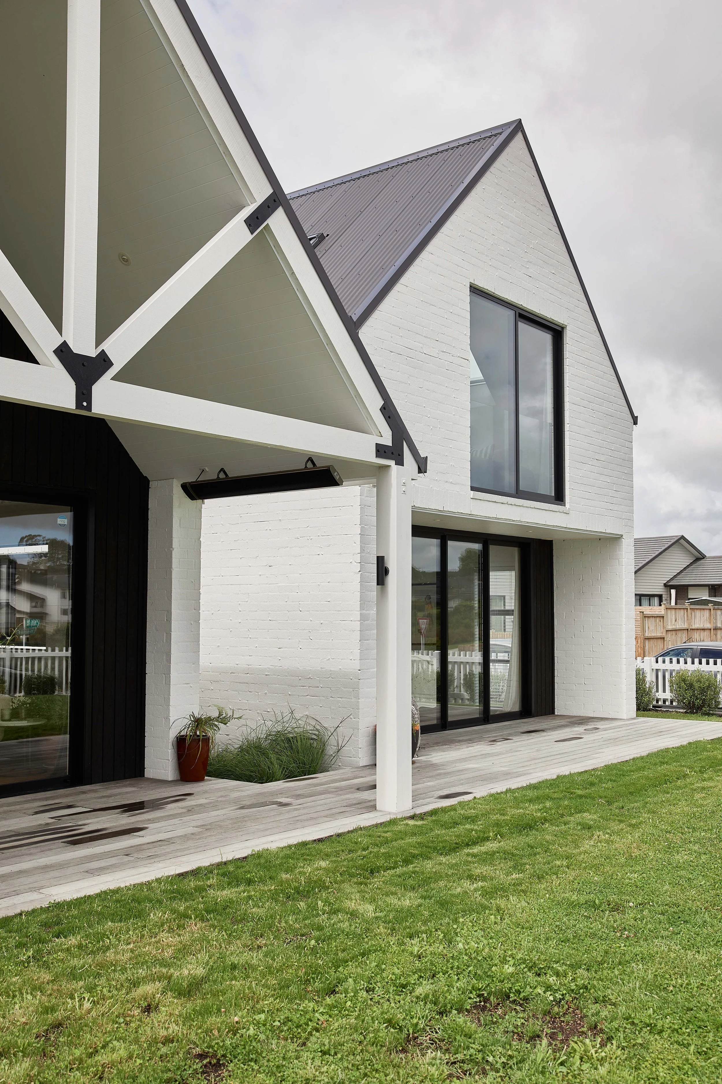 Modern two-story house with white brick exterior, large glass sliding doors, and a wooden deck. The house has a steep gabled roof with gray metal roofing and black trim. There are plants and grass in the front yard.