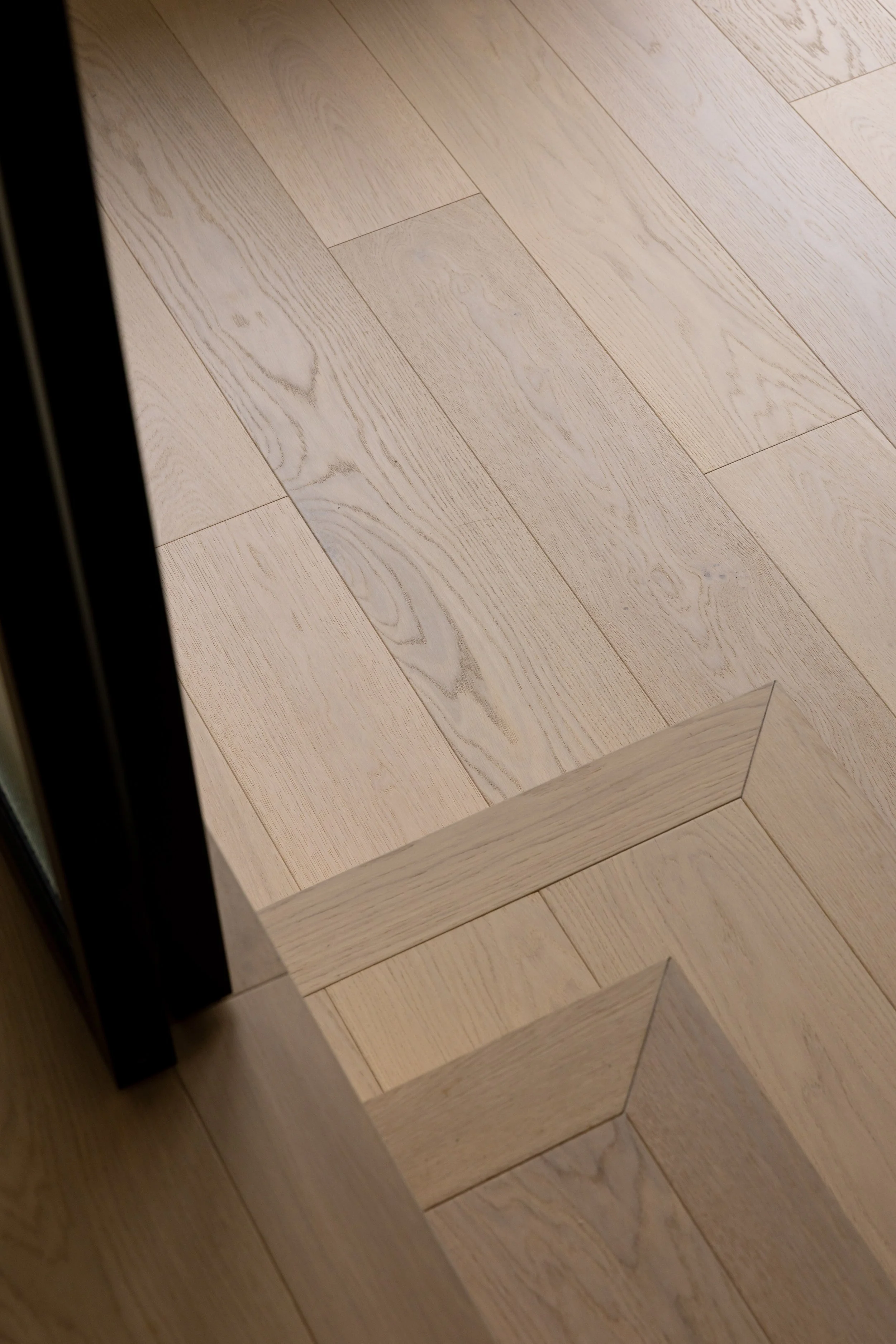 Close-up of a light-colored wooden floor with beveled edges, showing the corner of a door frame and part of a dark-colored furniture leg.