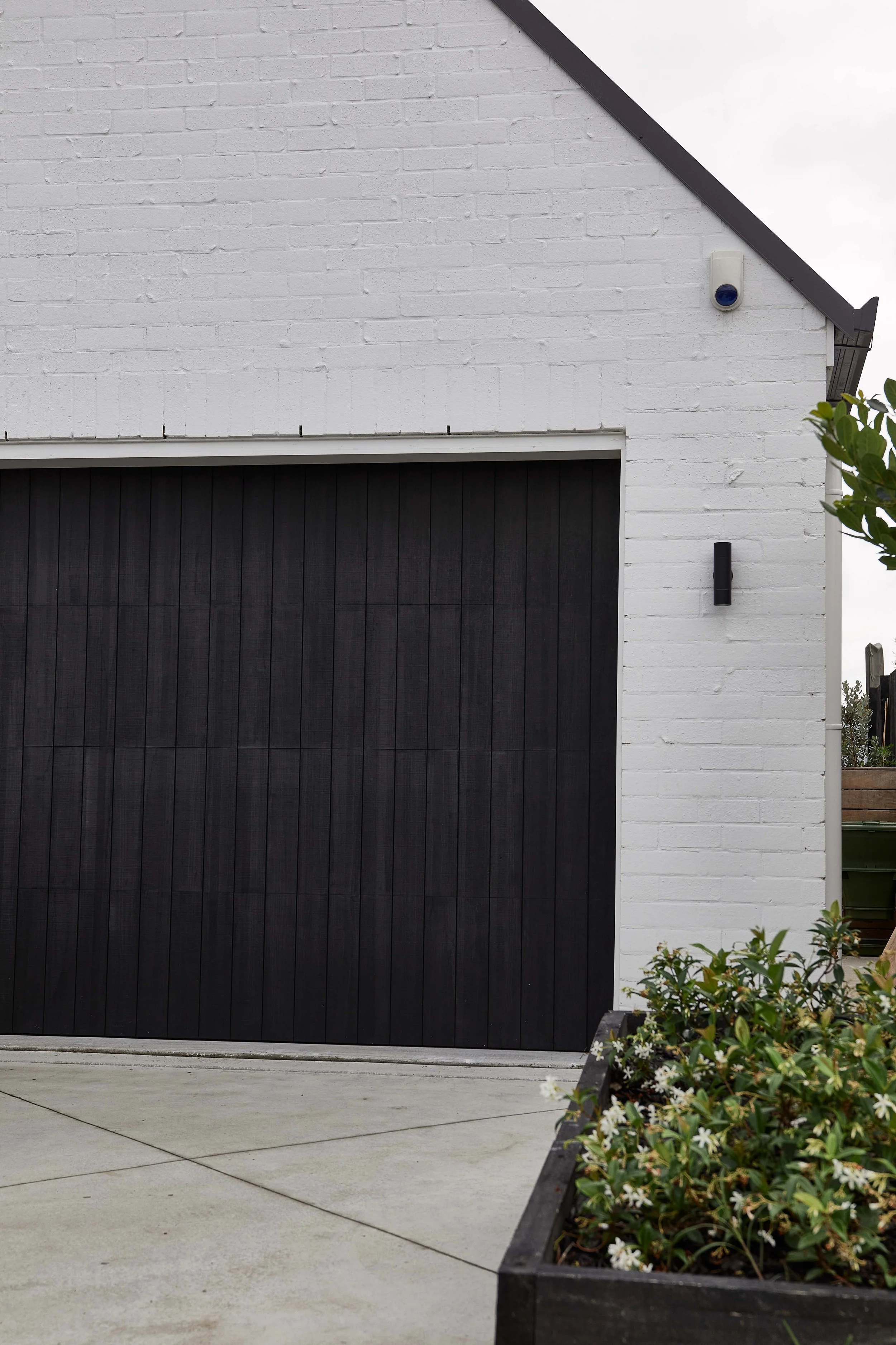 Close-up of a garage door and exterior wall of a modern house, with a bush and flower bed in foreground.