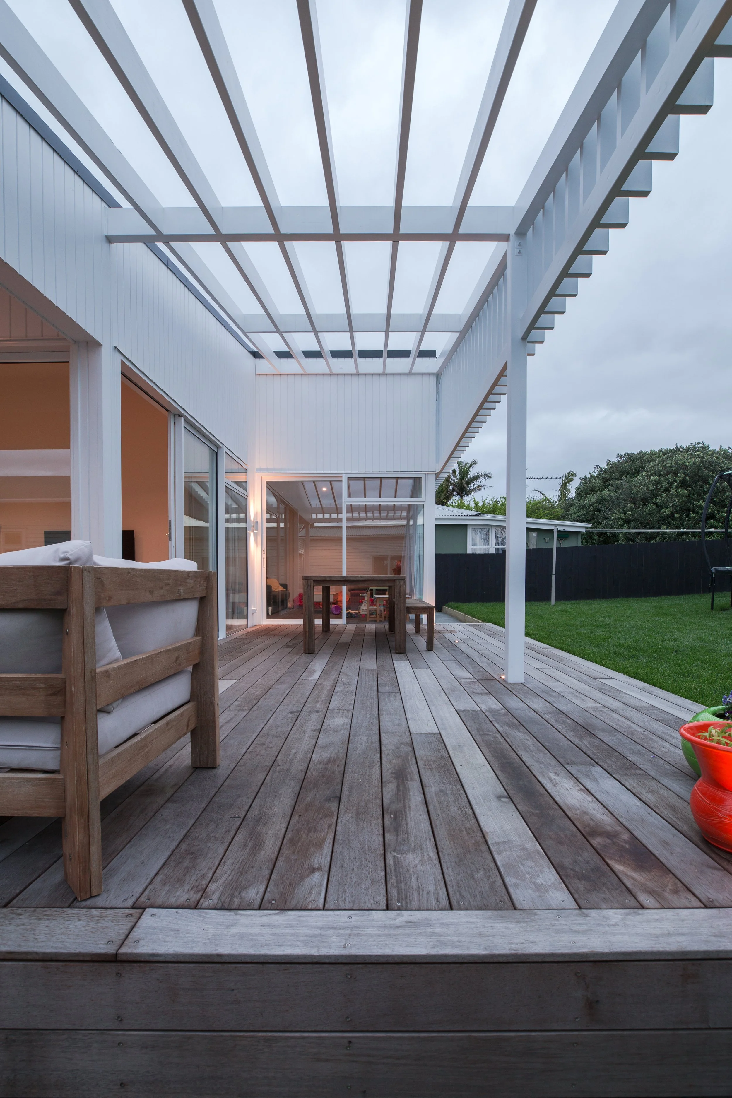 Outdoor patio with wooden decking, a dining table, and benches, attached to a modern white house with large sliding glass doors, a partially covered roof, and surrounding greenery.