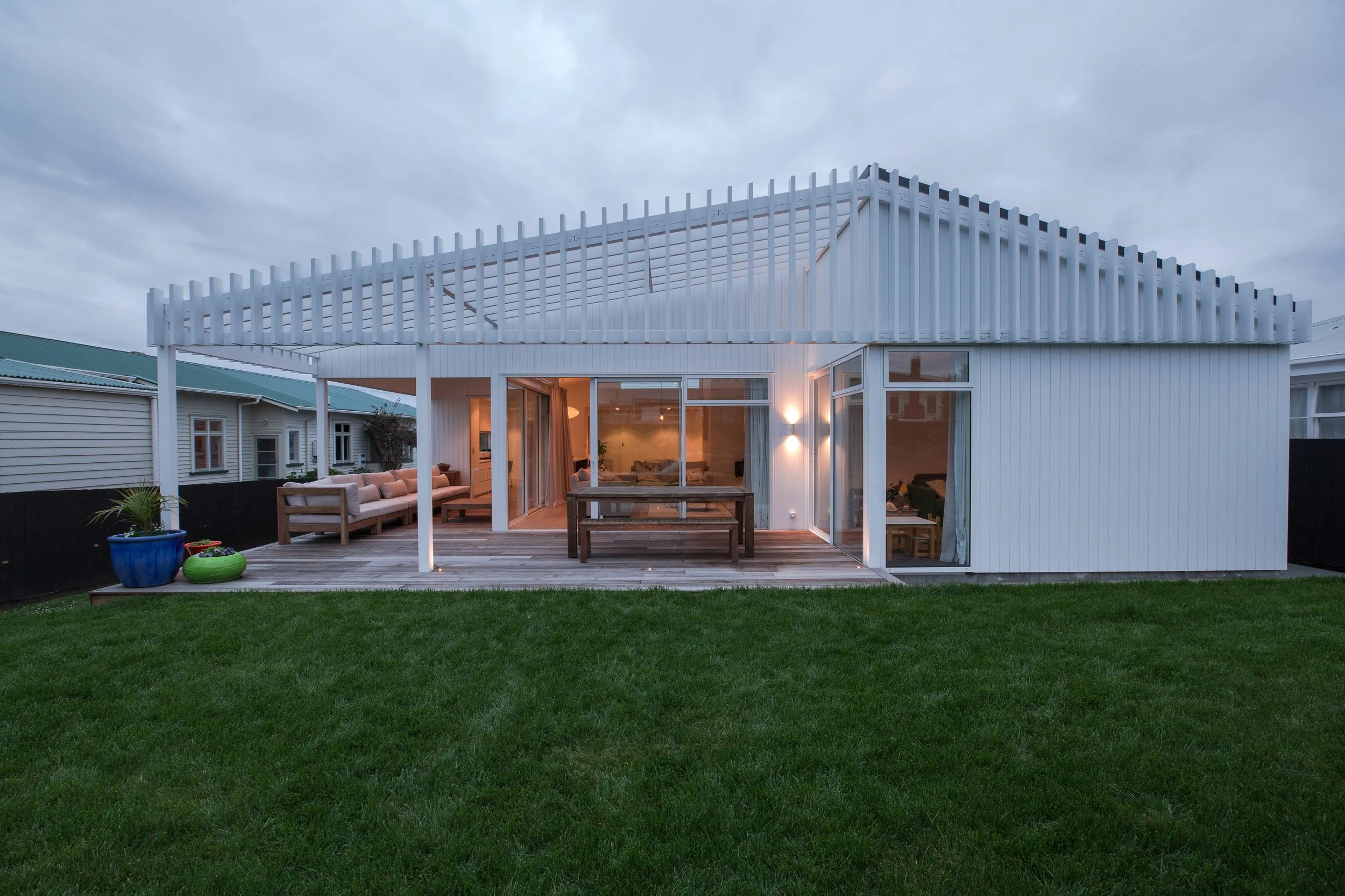 Modern white house with a wooden deck and outdoor seating, illuminated by exterior lighting, overlooking a green lawn under a cloudy sky.