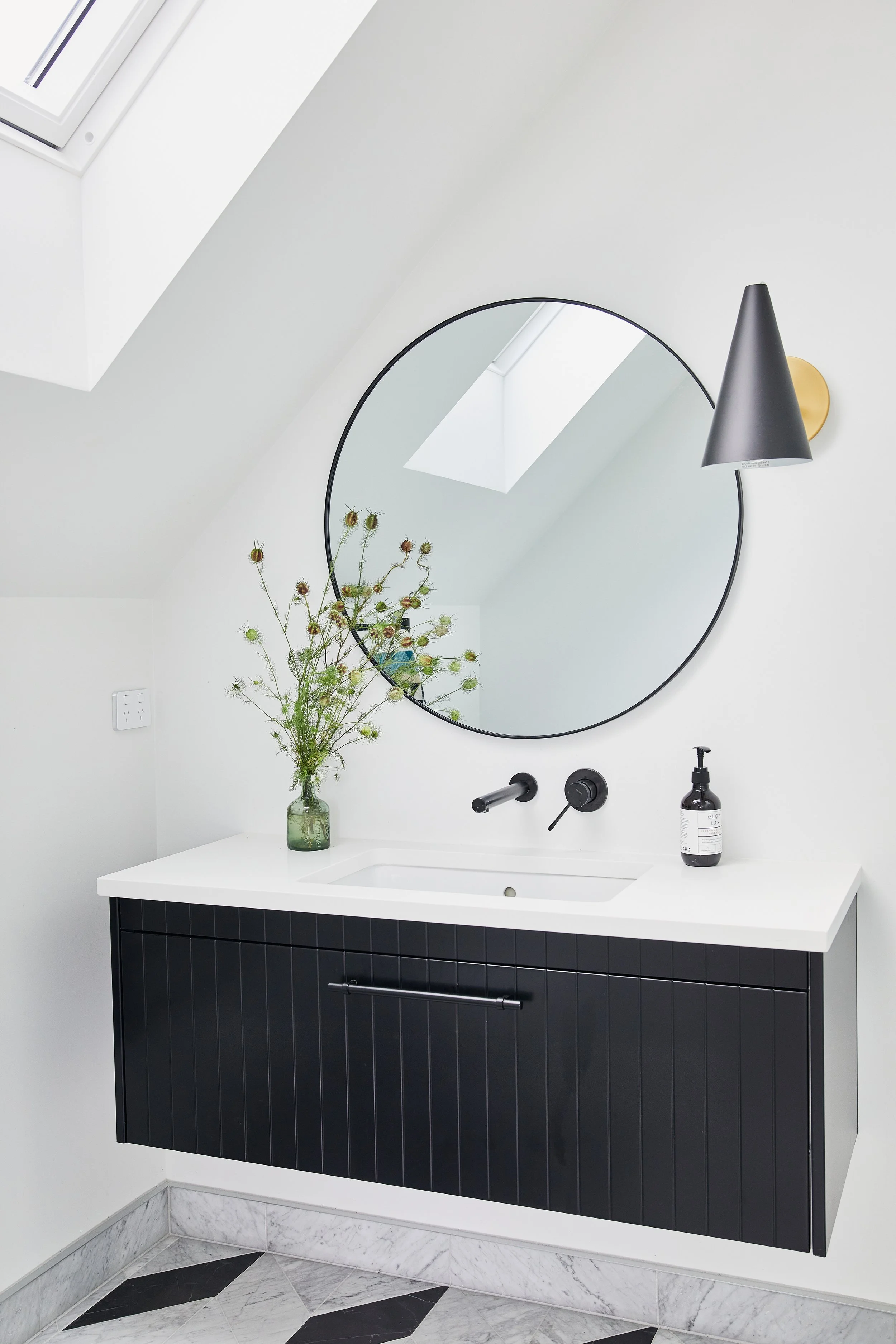 Modern bathroom vanity with a black cabinet, white countertop, round mirror, black wall-mounted faucet, black cone-shaped wall lamp, a vase with wildflowers, and a soap dispenser.
