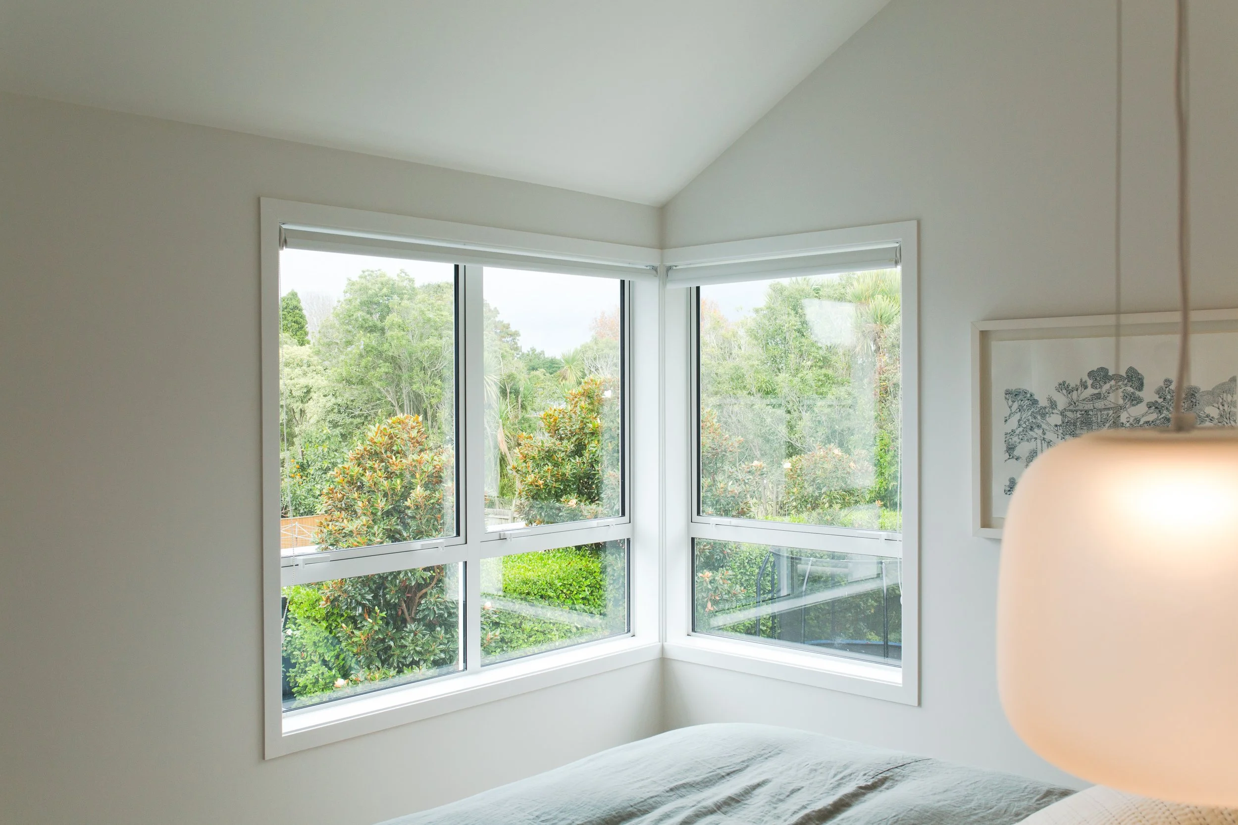 A bright bedroom with large corner windows overlooking a lush green garden.