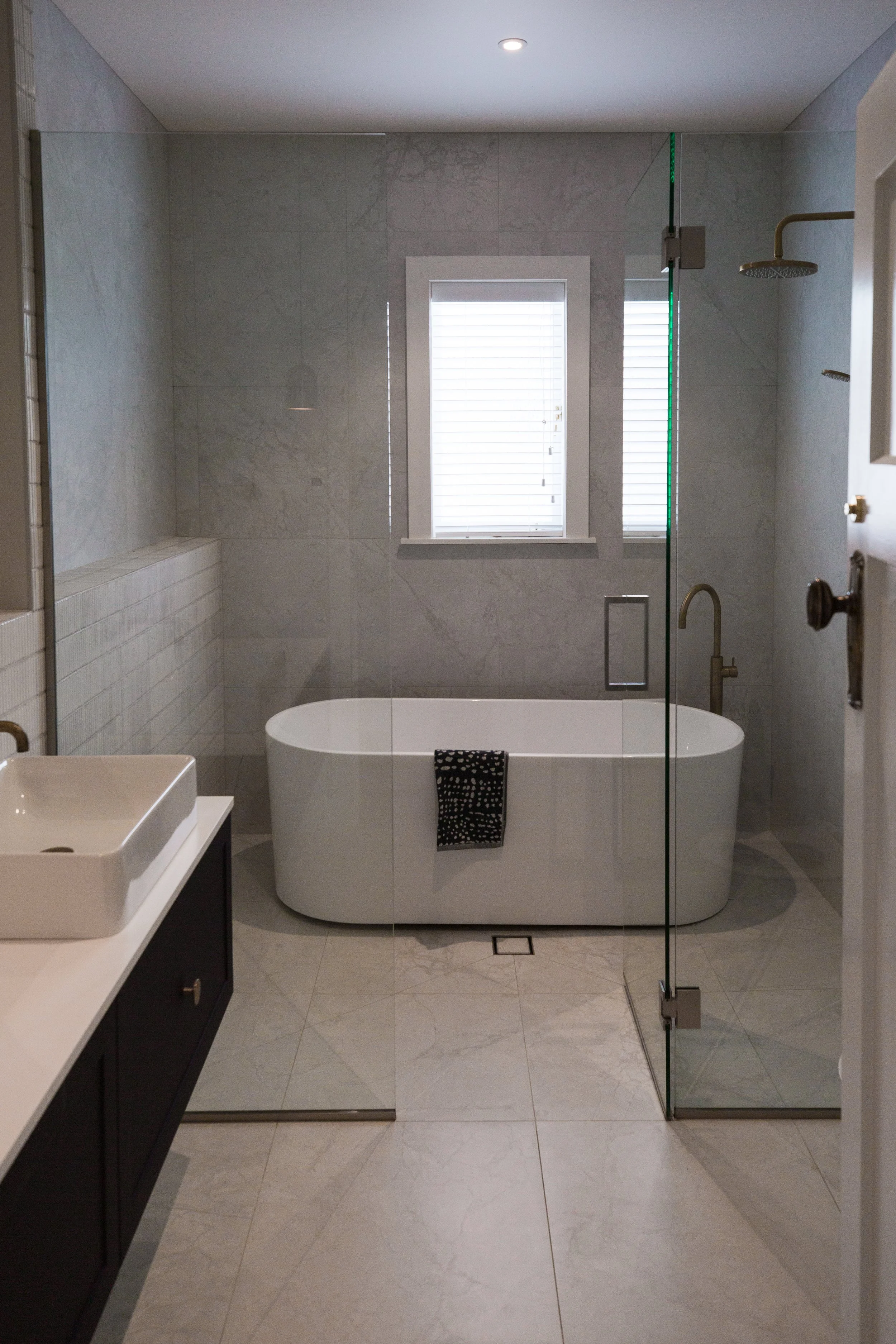 Modern bathroom with a freestanding bathtub, a glass shower enclosure, a window with white blinds, a wall-mounted faucet, and a dark vanity with a white vessel sink.
