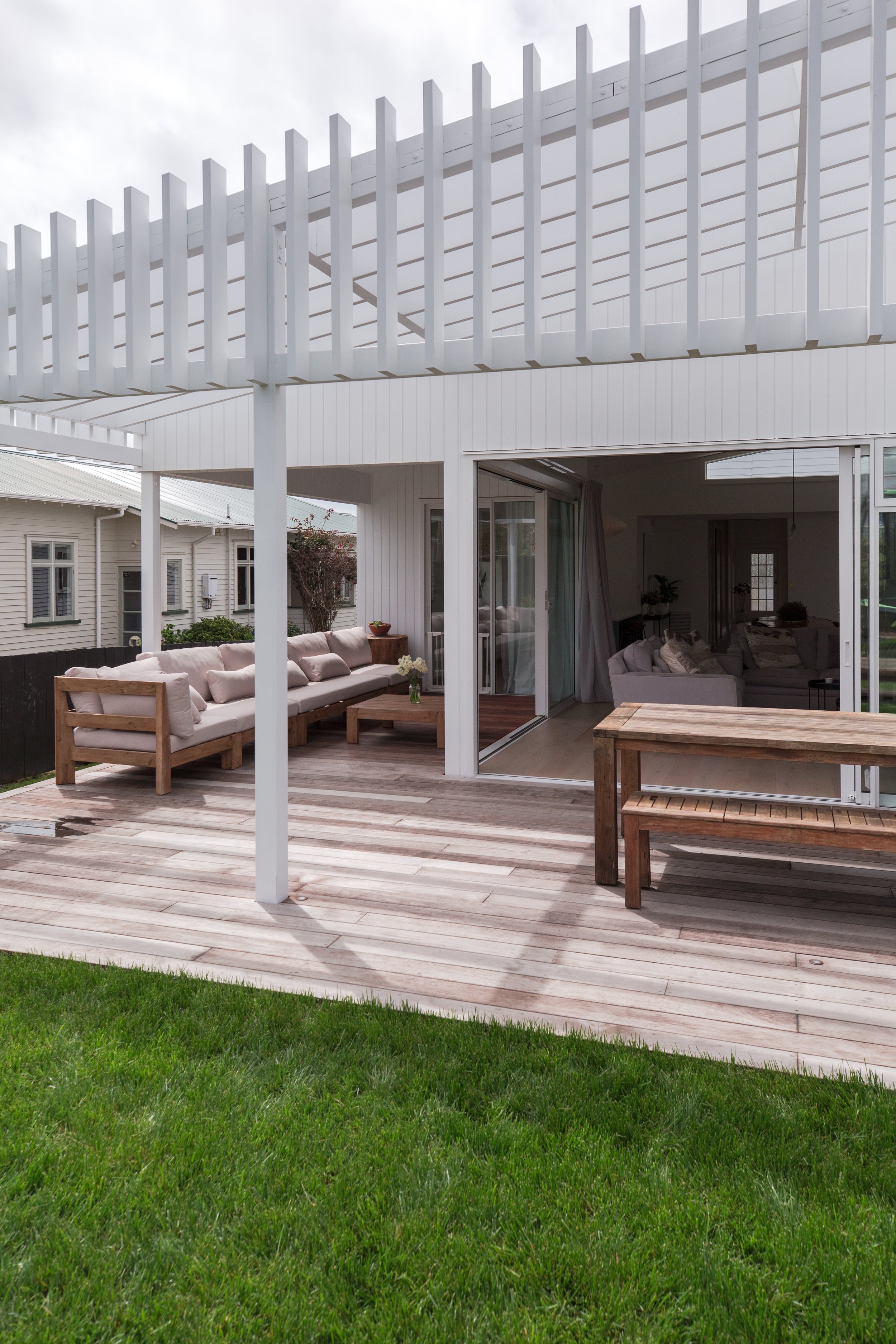Rear view of a modern house with a wooden deck, outdoor seating, and a pergola, overlooking a grassy yard.