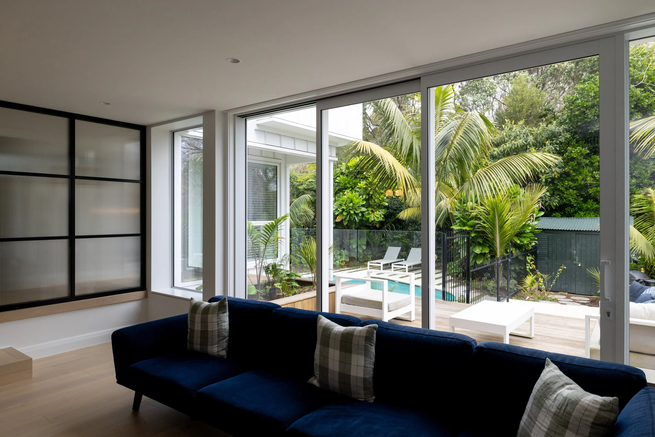 Living room with large sliding glass doors opening to a lush outdoor patio with lounge chairs and a small pool, decorated with green plants.